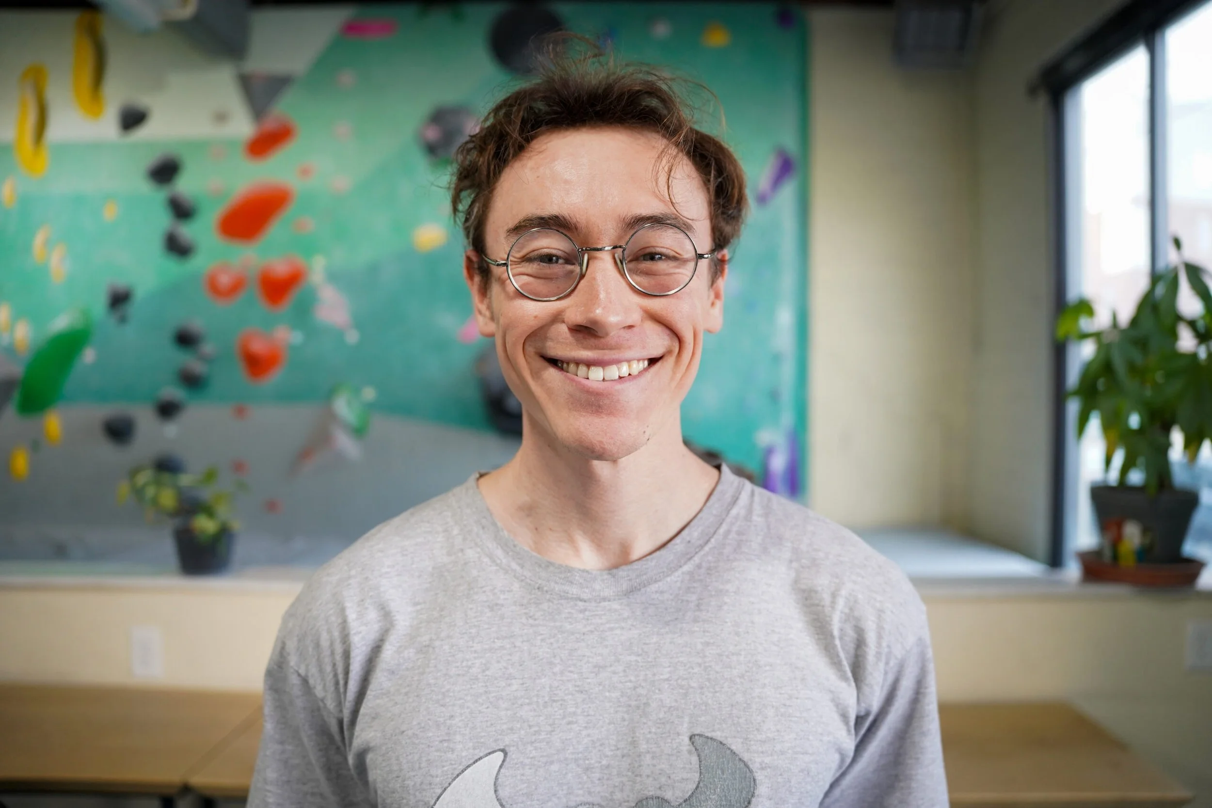 Smiling young man with glasses, wearing a light gray shirt, standing in front of a climbing wall with colorful holds and a window to the right, in an indoor gym.
