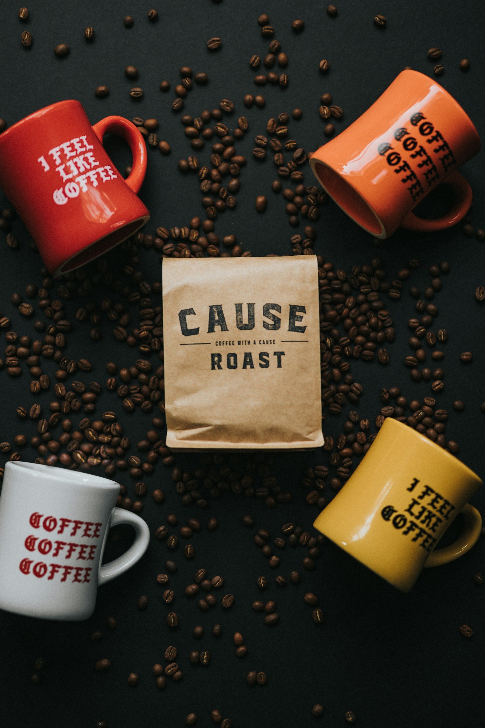Two coffee cups, one white and one orange, with the words "I feel like coffee" printed on them. Scattered coffee beans surround the cups, and a brown paper bag with "Cause Roast" written on it is in the center.