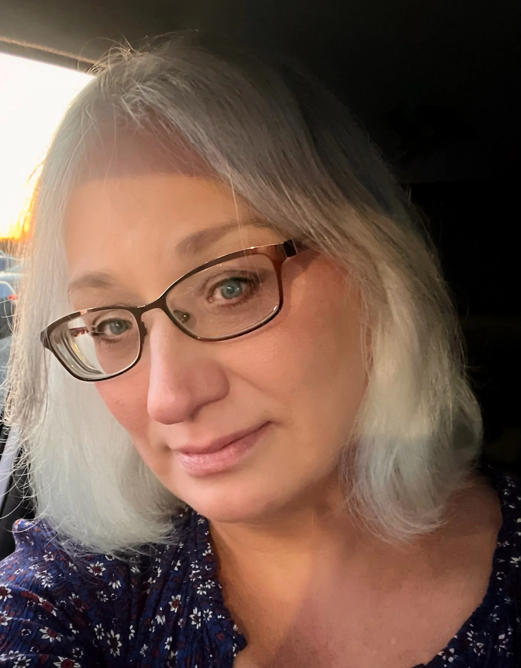 Close-up of a woman with gray hair, glasses, and a smile, wearing a dark floral top.