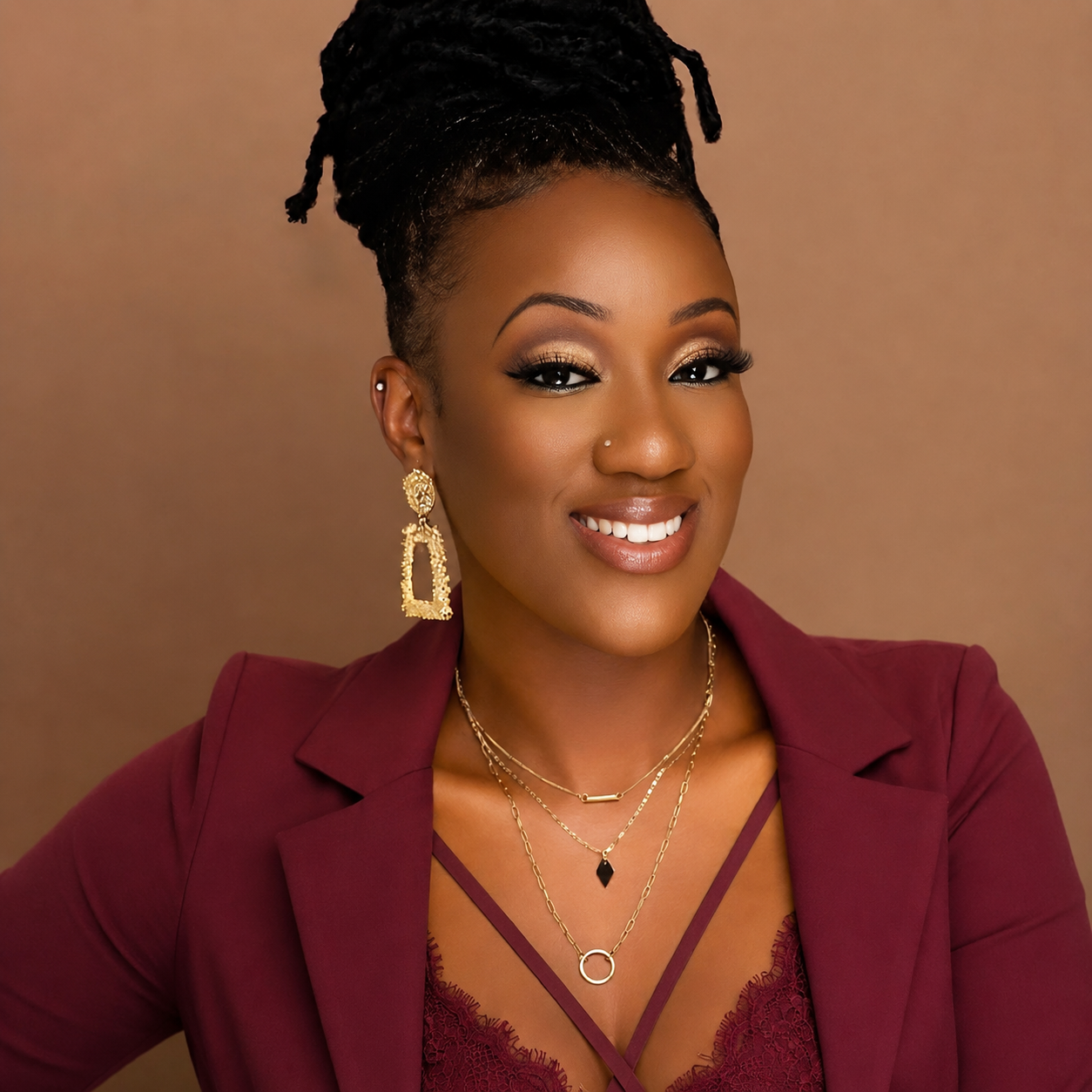 A woman smiling with gold jewelry, including earrings, necklaces, and a nose ring, wearing a burgundy blazer and lace top, with a neutral background.