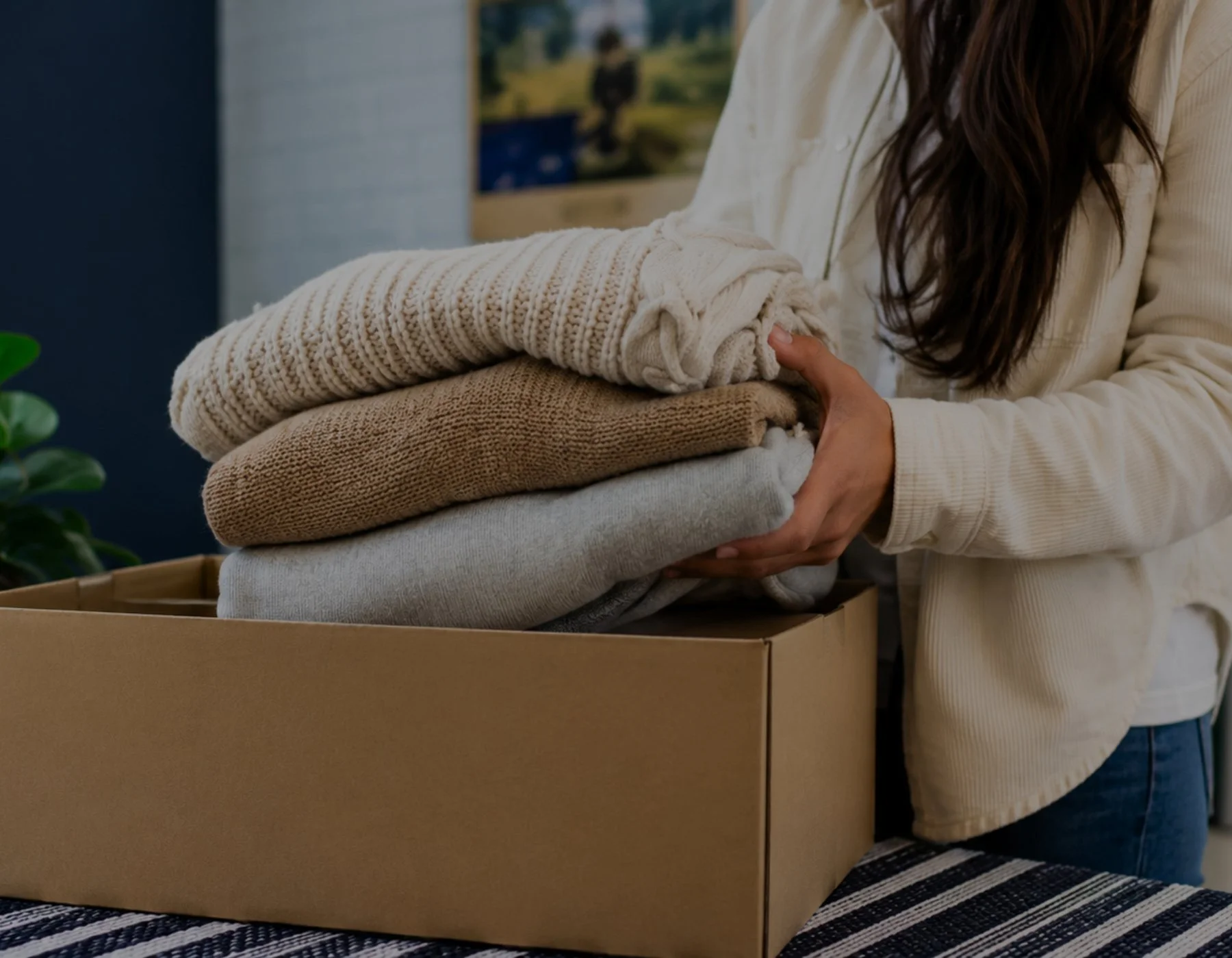 A person holding a box of folded sweaters, ready to take to clothing recycling bins at Home+ McGraths Hill.