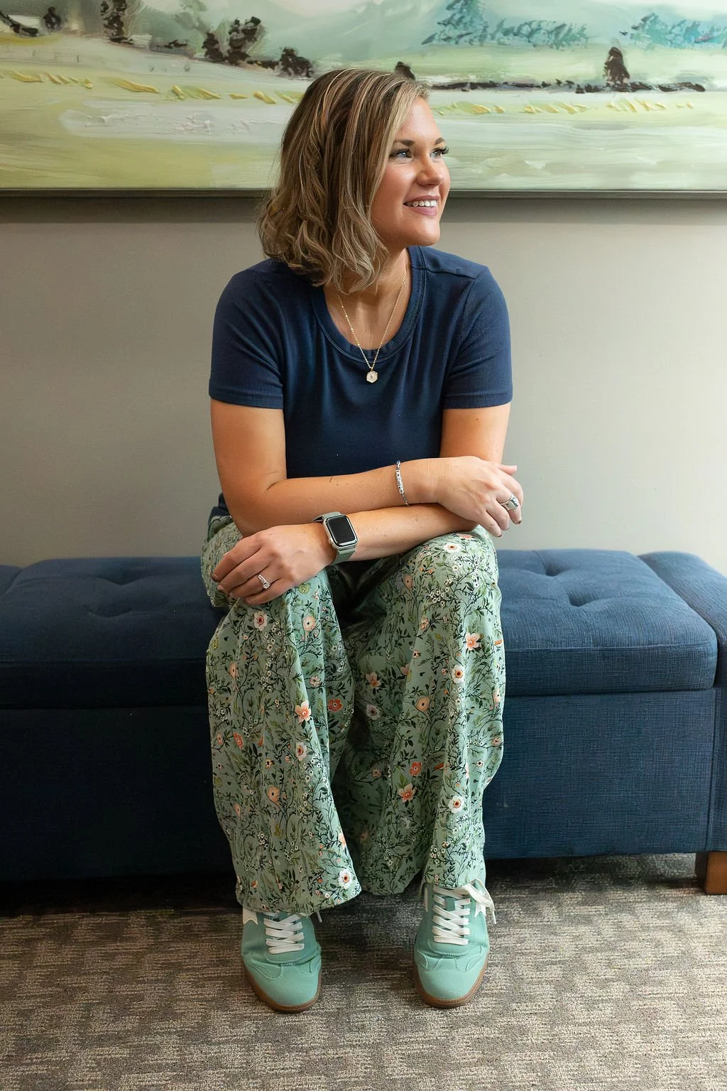 A woman with shoulder-length wavy hair sitting on a blue couch, wearing a navy T-shirt, floral pants, mint green sneakers, and a smartwatch, smiling and looking to her left with a framed abstract painting in the background.