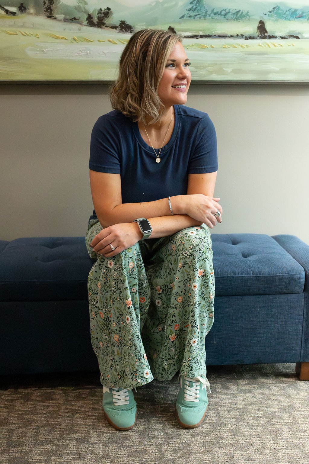 Christian mental health speaker in Sioux Falls, SD, Krysta Winter of Christian Family Care, smiling and gazing off to her right while seated on a blue bench in her counseling office, wearing a navy top and floral pants.