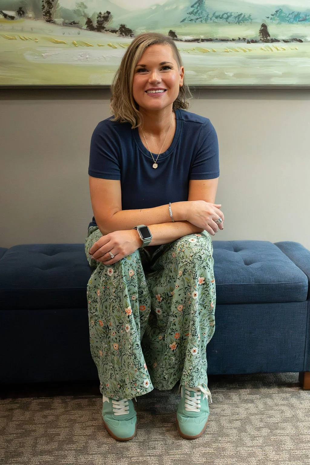 Portrait of Krysta Winter smiling while seated on a blue bench, wearing a navy shirt, floral pants, and mint green sneakers.