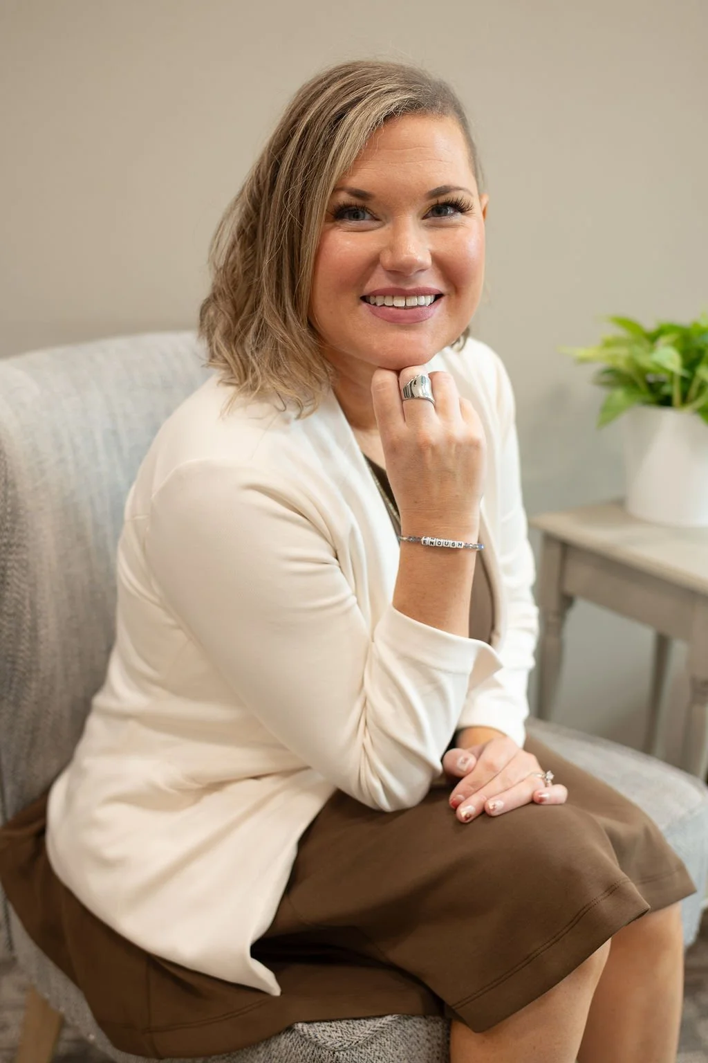 Christian mental health speaker in Sioux Falls, SD, Krysta Winter of Christian Family Care, smiling at the camera while seated in an upholstered chair, wearing a white blazer and brown dress with her hand resting beneath her chin.