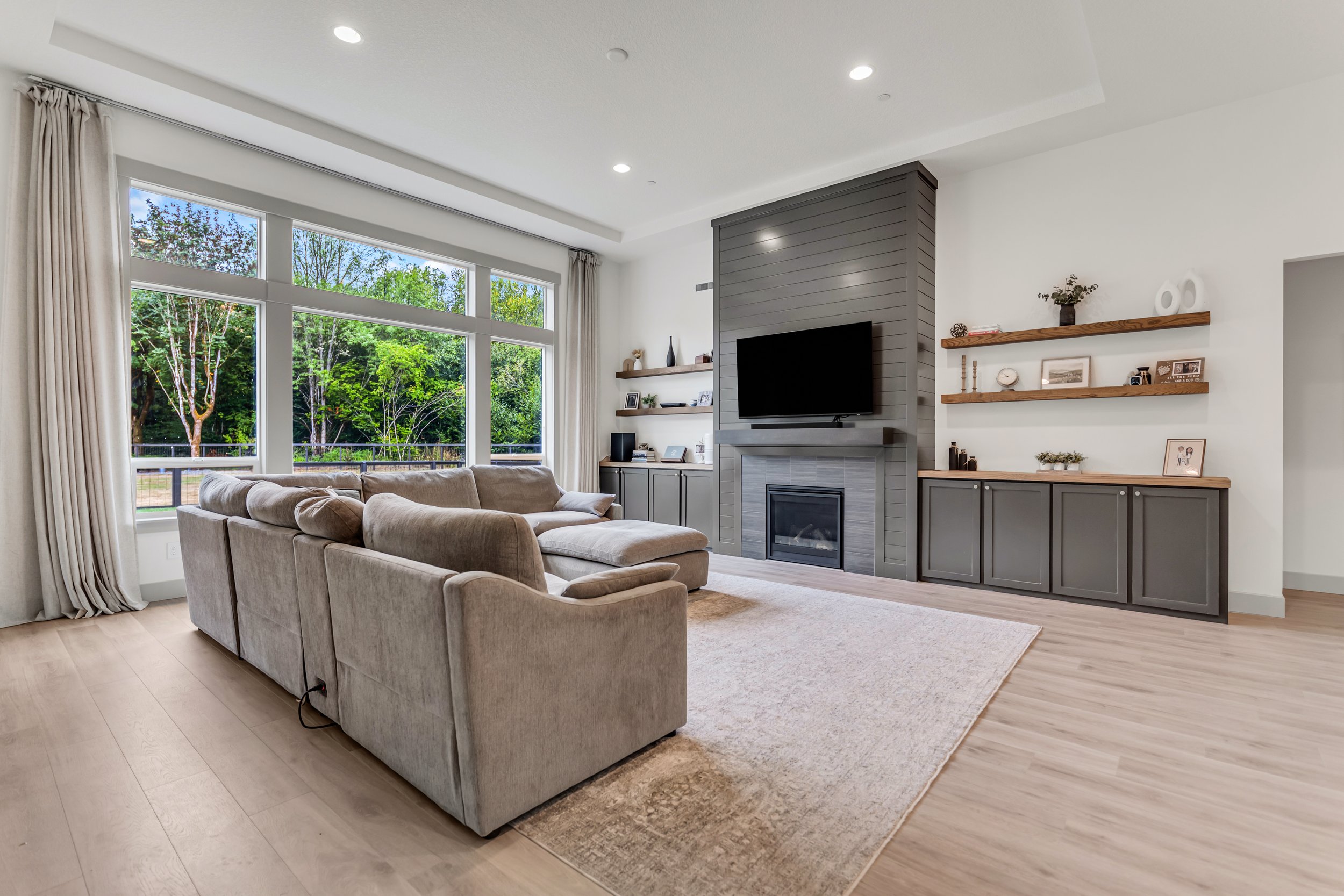 Bright living room with beige sectional sofa near a large window, gray accent wall with mounted TV, and wooden shelves with decor, overlooking trees outside.