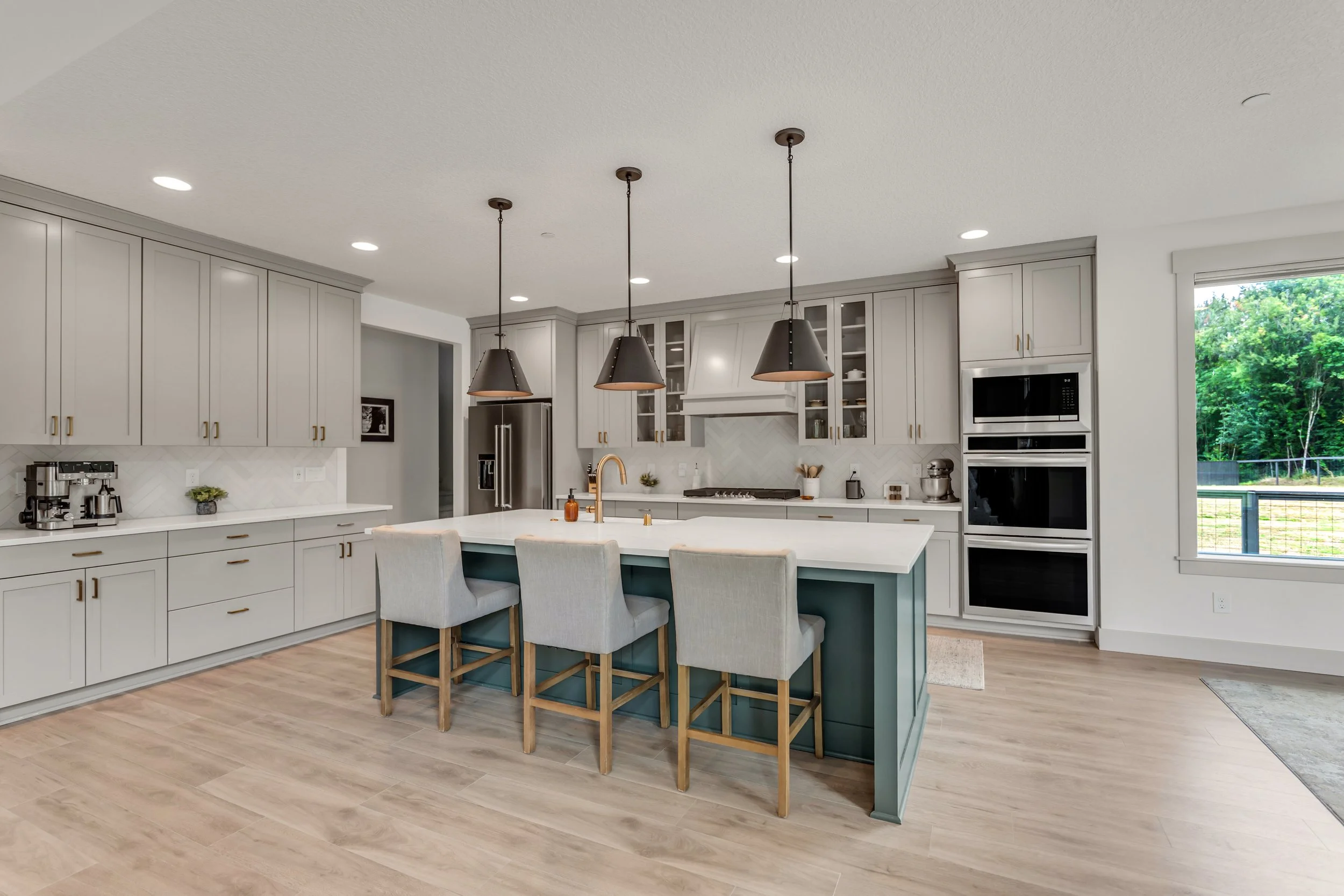 Modern kitchen with white cabinets, a large island with bar seating, pendant lights, stainless steel appliances, and a window showing greenery outside.