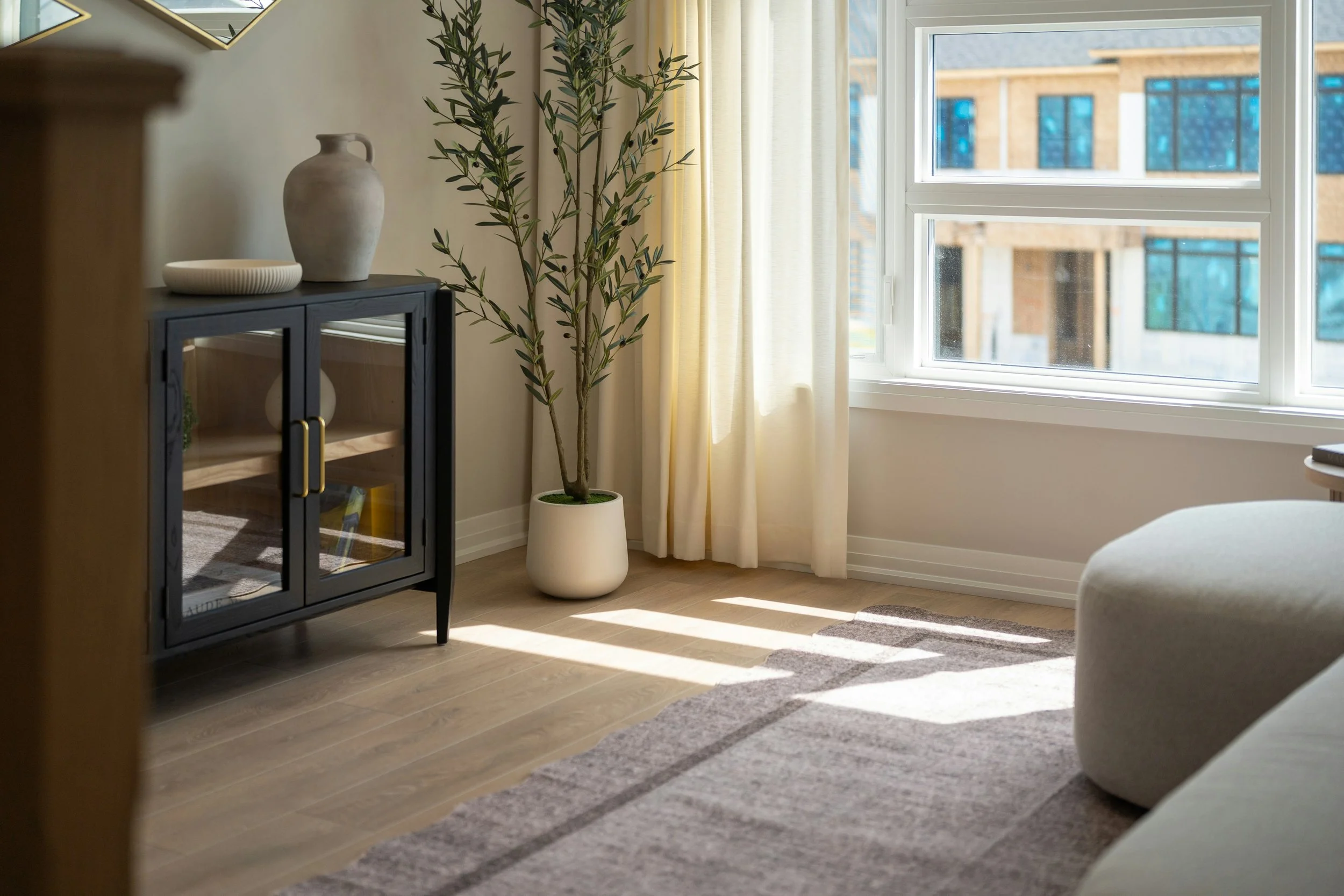 A cozy living room corner with a potted plant next to a large window with yellow curtains, a black cabinet with decorative vases and bowls, and sunlight streaming in.