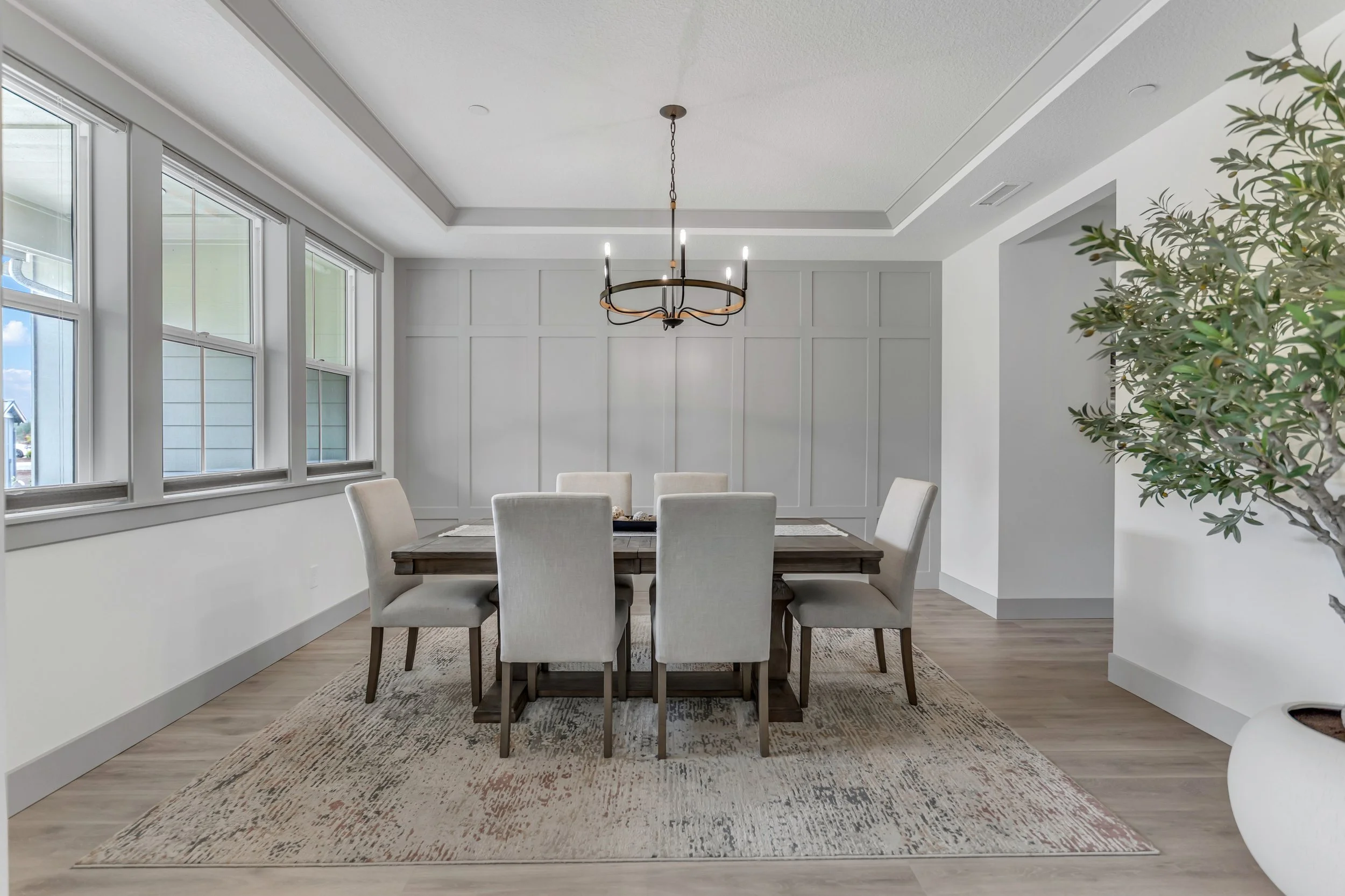 A dining room with a wooden table surrounded by six light-colored upholstered chairs. Large windows on the left wall allow natural light into the room. There is a decorative chandelier hanging from the ceiling, and a rug under the table. A large leaf