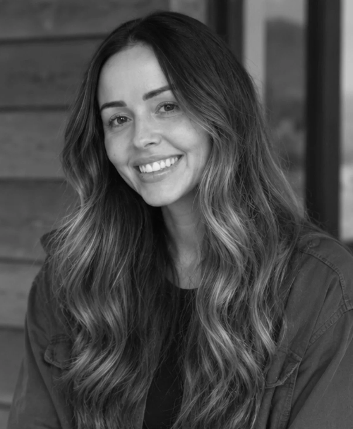 A black and white portrait of a smiling woman with long wavy hair, wearing a dark shirt or jacket, in front of a wooden background.