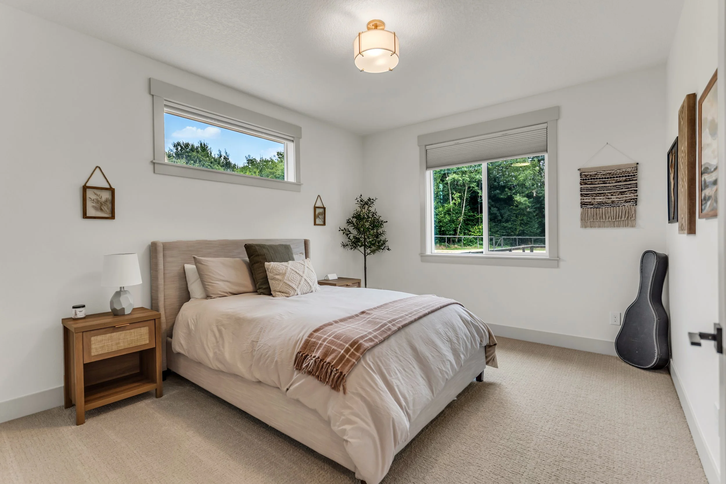 A bedroom with a bed, nightstand, and decorative wall art. Two windows reveal a view of green trees outside, and a guitar leans against the wall near a door.