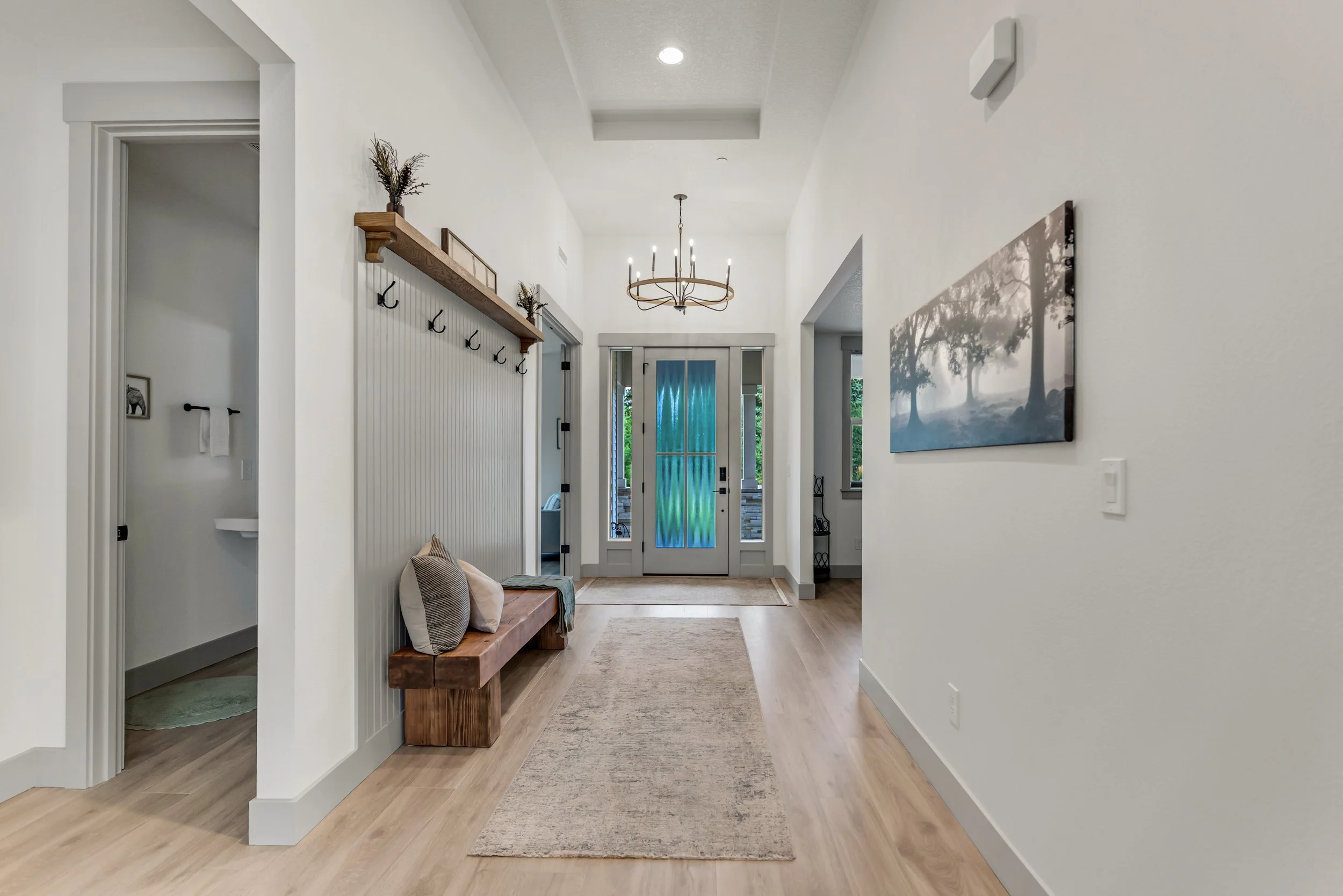 A bright, modern entryway with light wood floors, a beige runner rug, a wooden bench with pillows, a framed artwork of trees on the wall, a chandelier, a glass front door, and a narrow side window. There's a wall with hooks and shelves for decor and 