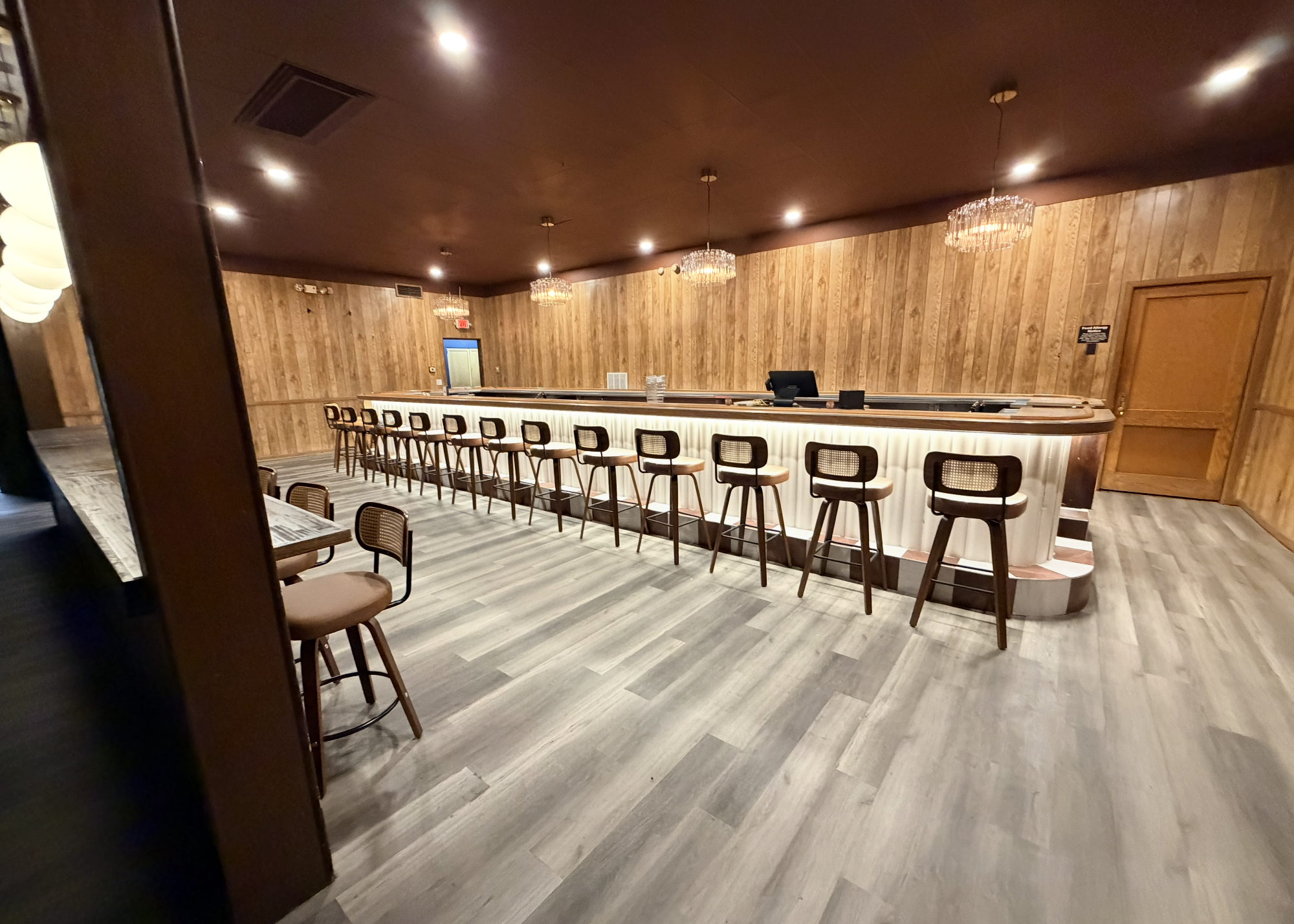 Empty bar area with a curved counter, bar stools, wooden paneled walls, chandeliers, and a doorway in the background.