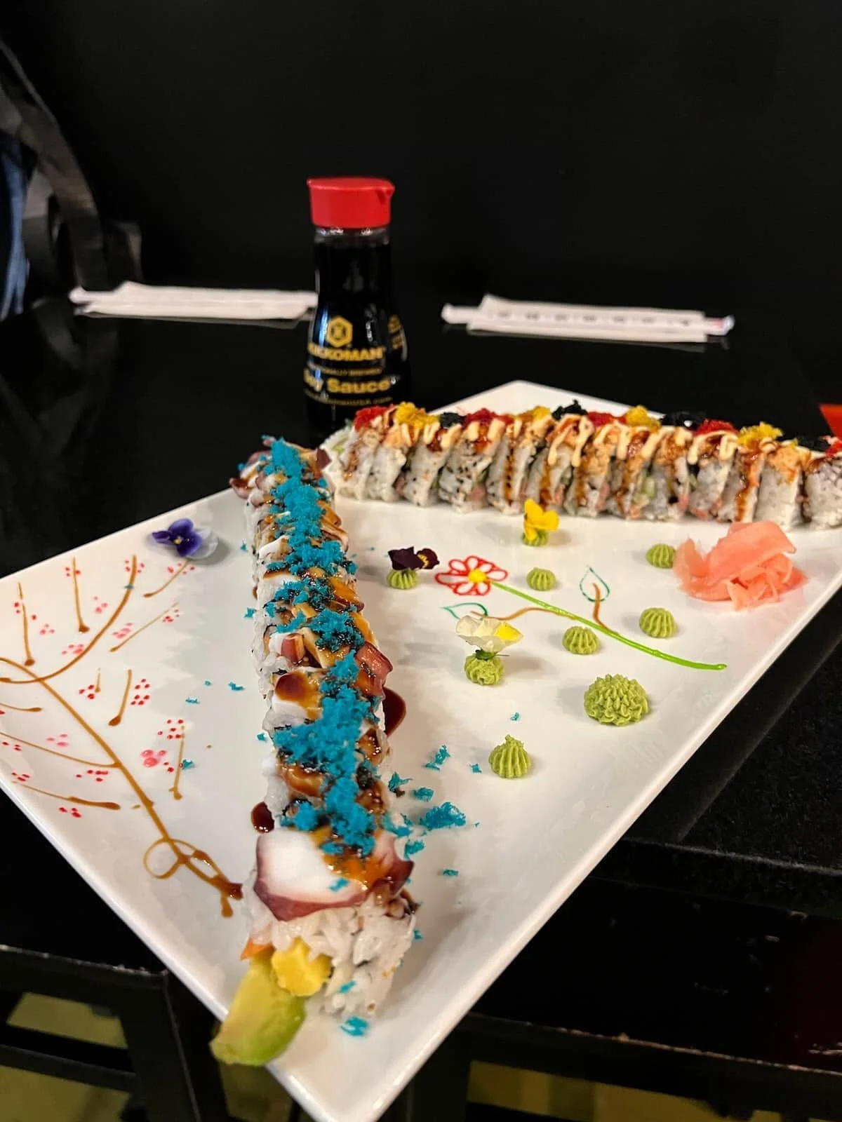 A decorative sushi platter with sushi rolls topped with sauce and blue sprinkles. The plate has floral and branch designs with small green, yellow, purple, and toasted toppings, along with pink pickled ginger and wasabi. In the background, there is a soy sauce bottle and some utensils.