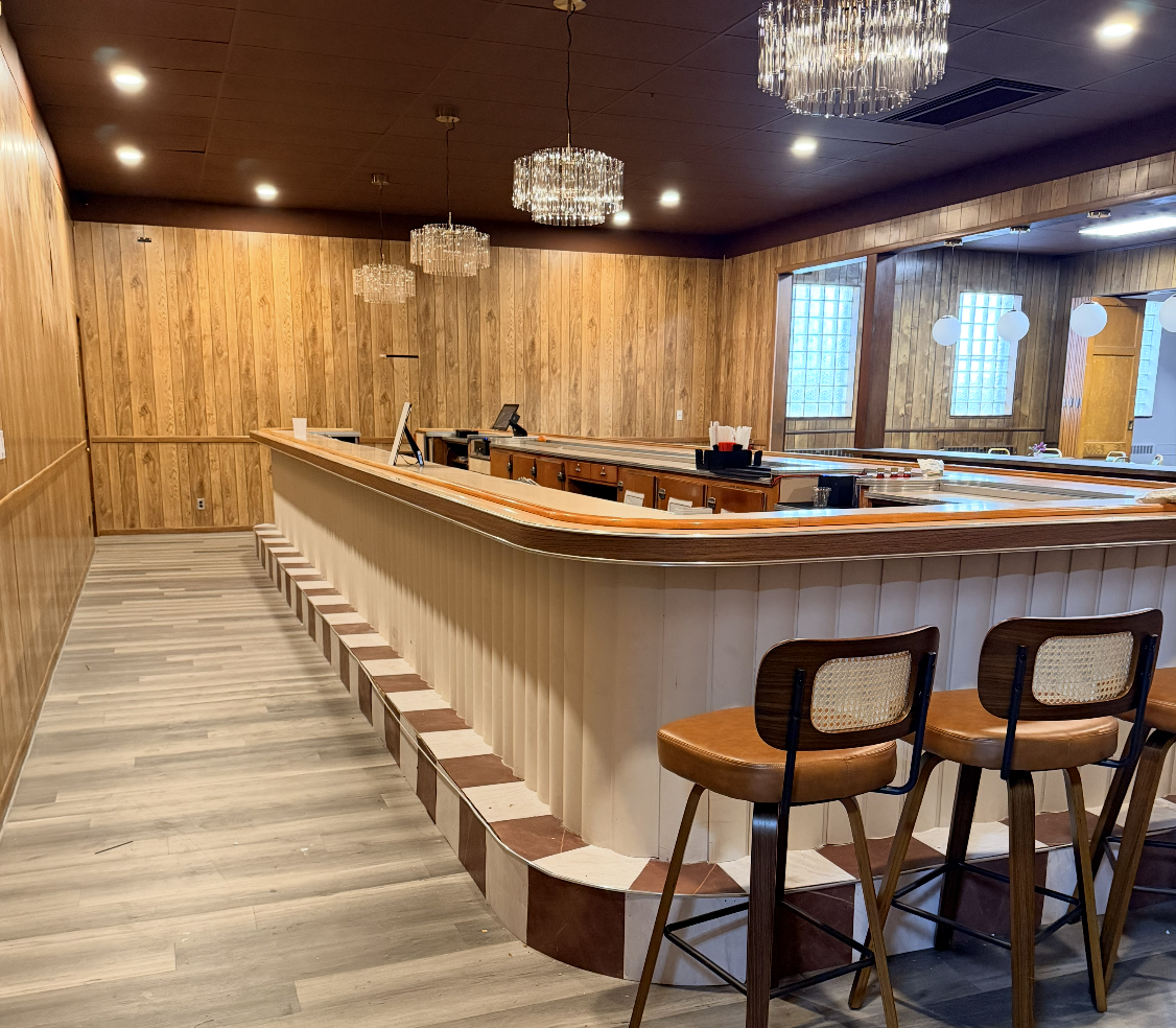 Empty bar area with wood paneling, chandeliers, stools, and a counter in a cozy restaurant or pub.