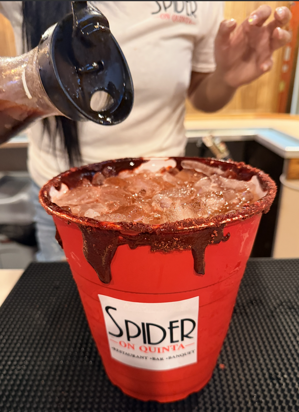 A person pouring a dark beverage into a red bucket labeled 'Spider on Quinta' at a restaurant or bar.