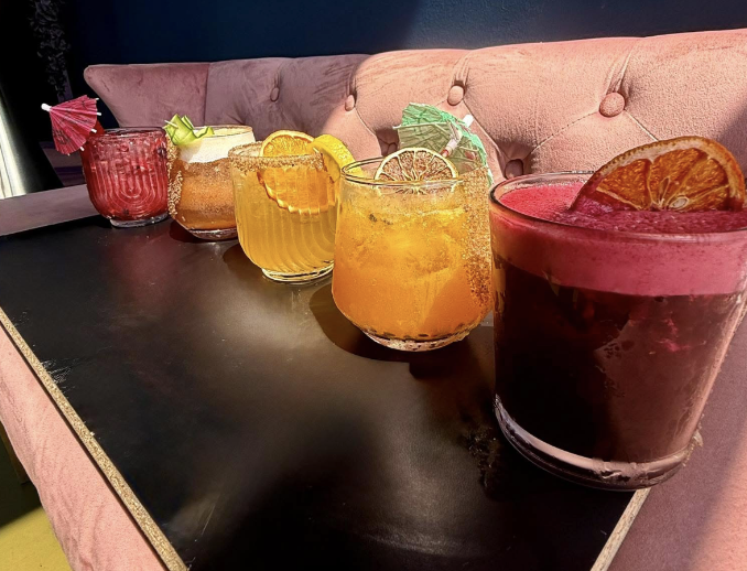 Six colorful cocktails with garnishes on a dark table in a restaurant or bar setting.