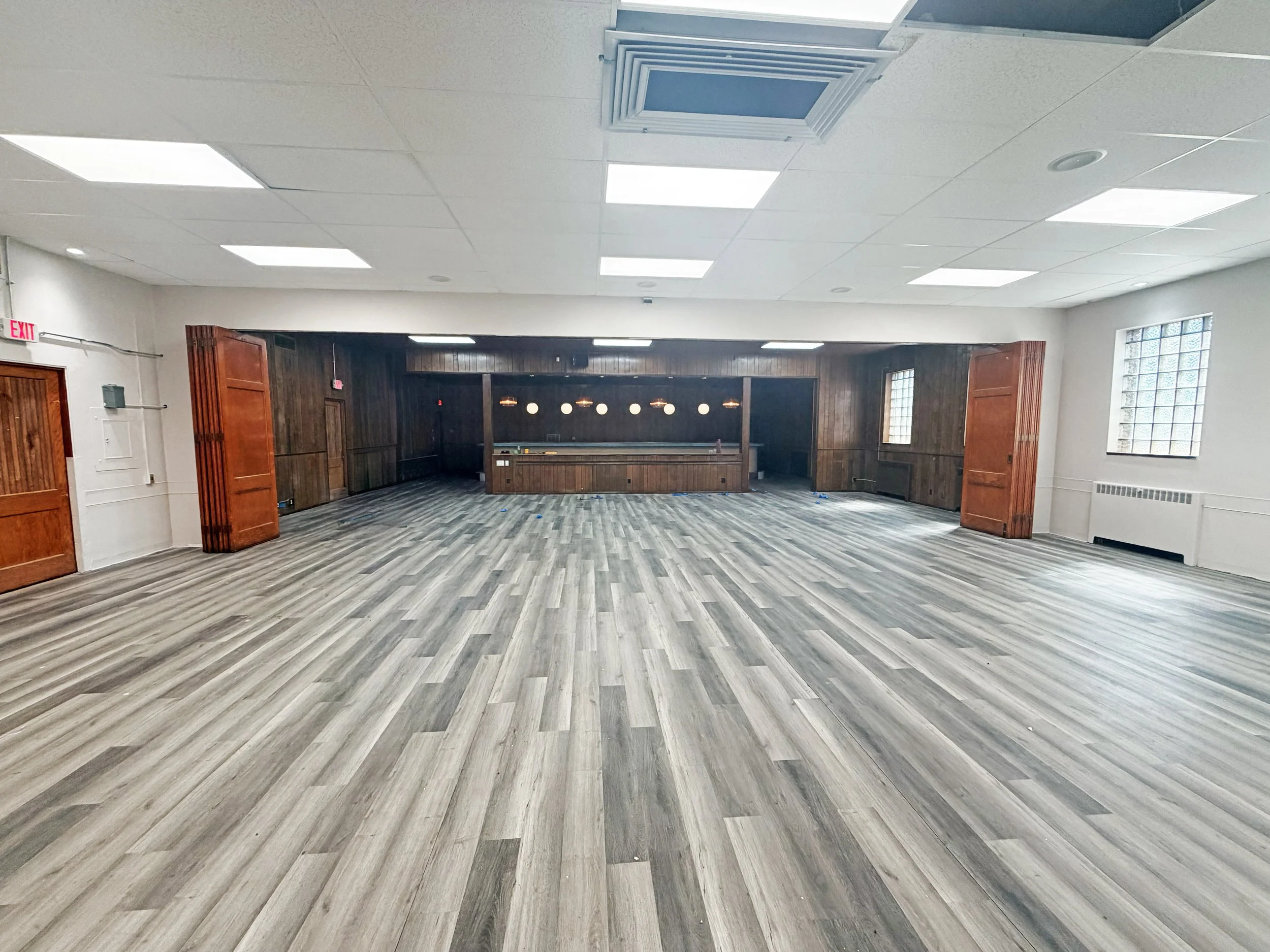 Empty room with wood-paneled walls, large windows, and a wood bar at the back. The floor is gray wood, and the ceiling has bright square lights.