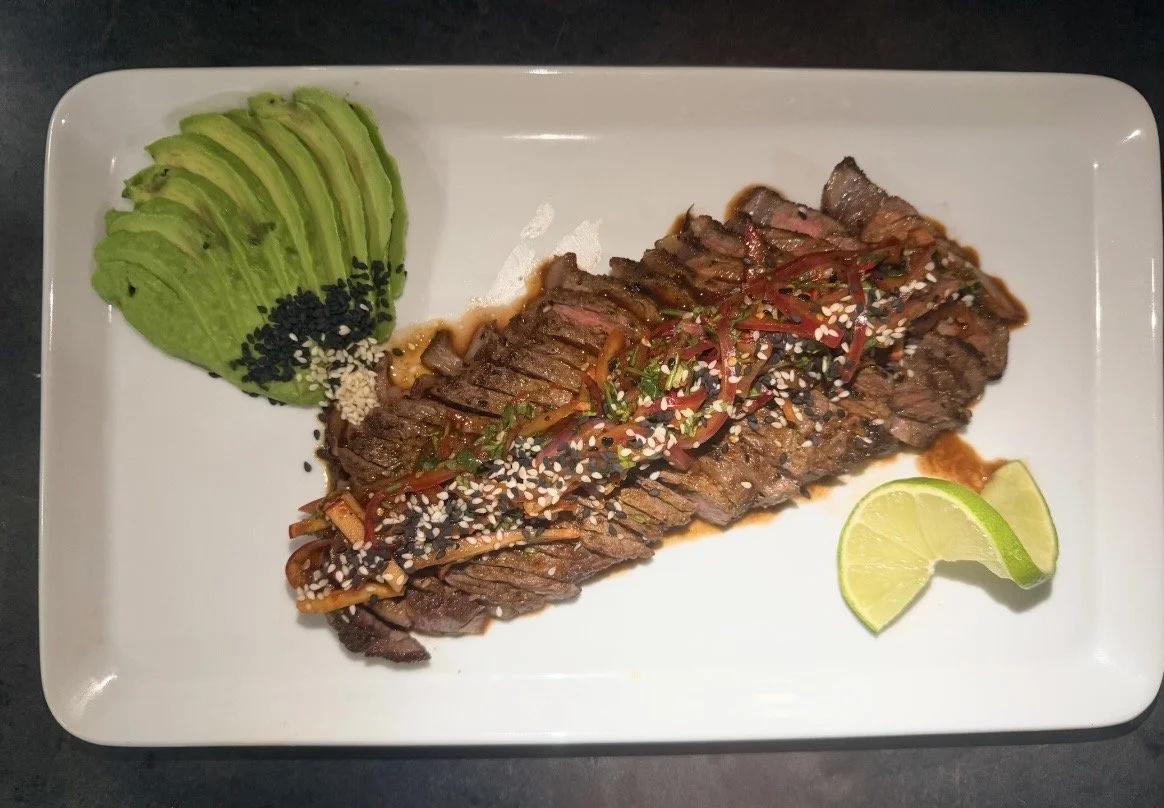 Sliced steak topped with sauce, sesame seeds, and sliced red peppers, served with sliced avocado sprinkled with black sesame seeds and two lime wedges.