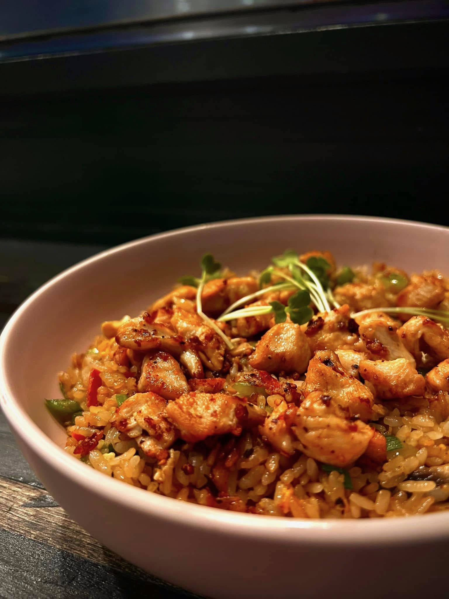 Bowl of fried rice with chicken and green garnish, on a wooden surface.