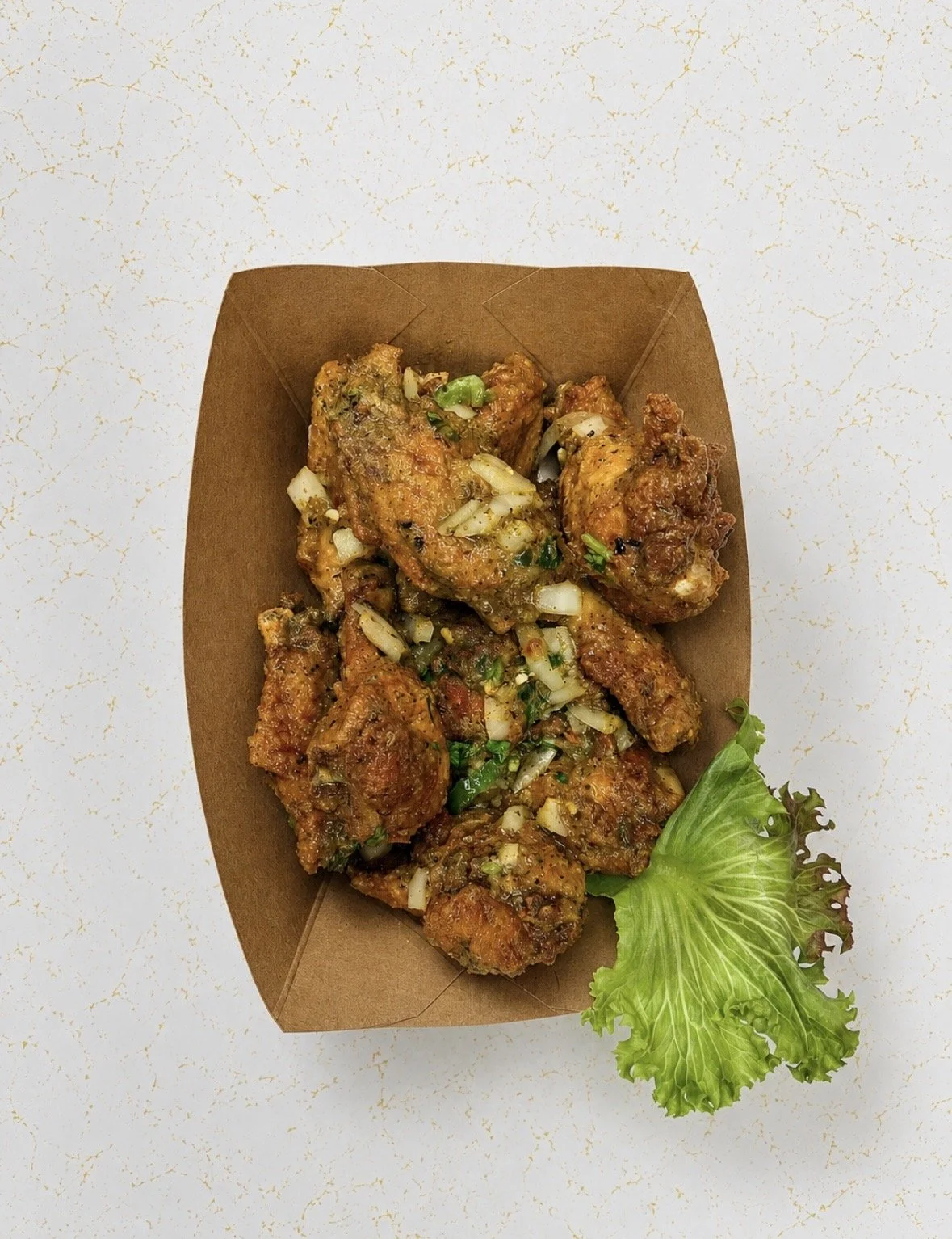 Fried chicken wings garnished with chopped green onions and served with a leaf of lettuce in a brown takeout container on a white speckled surface.