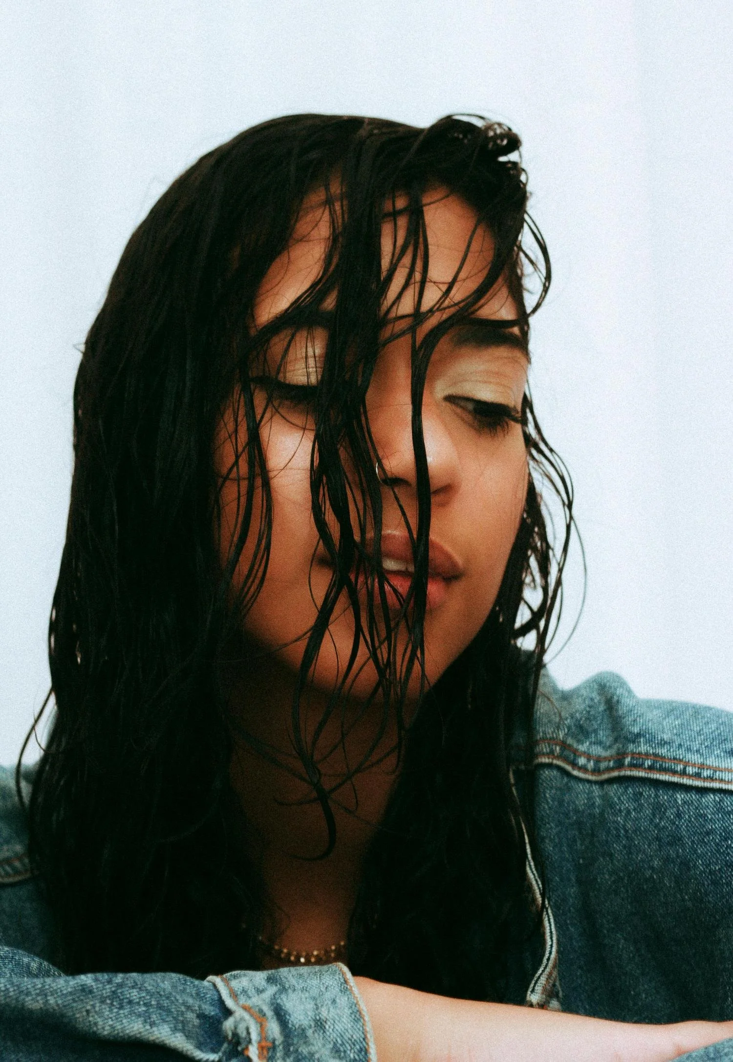 Close-up of a woman with wet black hair, closed eyes, and a calm expression, wearing a denim jacket and a pearl necklace.