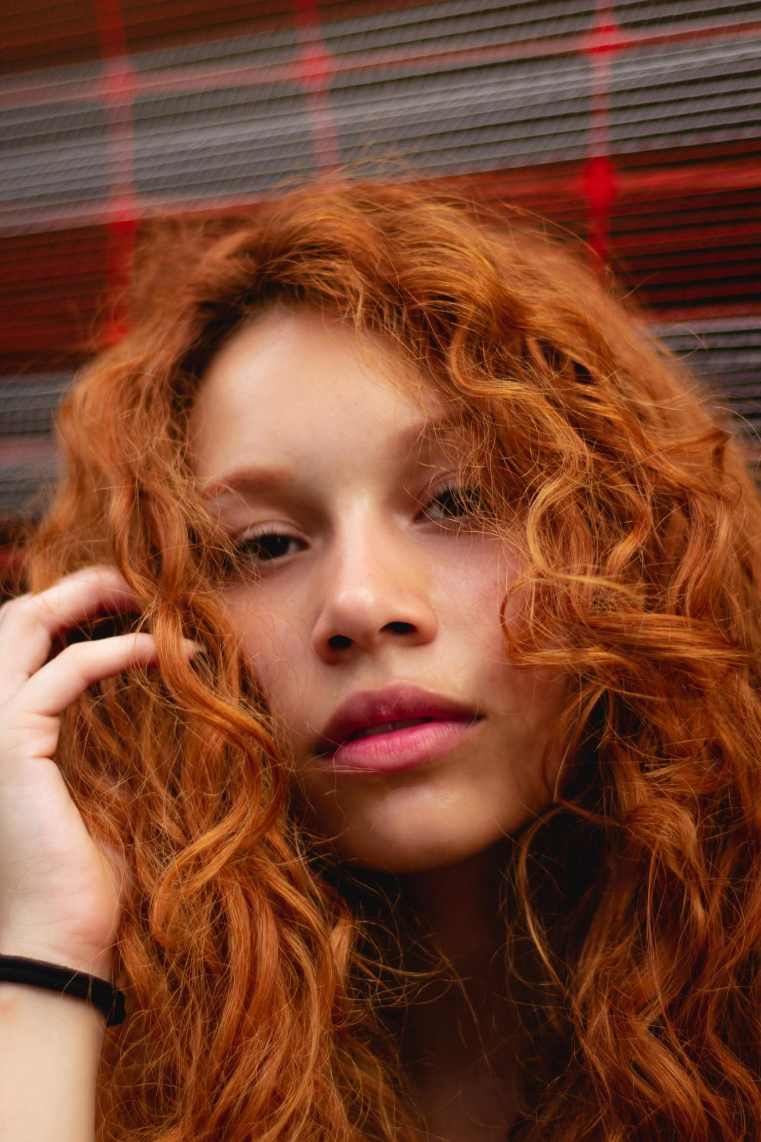 Close-up of a young woman with long, curly red hair and light skin, touching her hair with her hand, standing in front of a red and gray background.