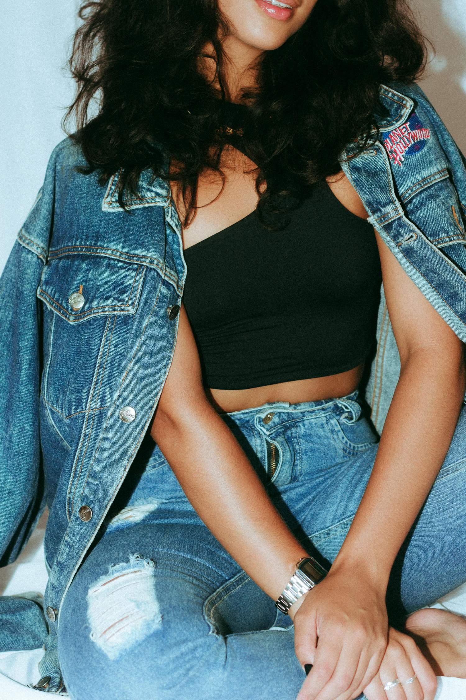 Close-up of a woman wearing ripped blue jeans, a black top, and a denim jacket with a "Planet Hollywood" patch, sitting with hands clasped.
