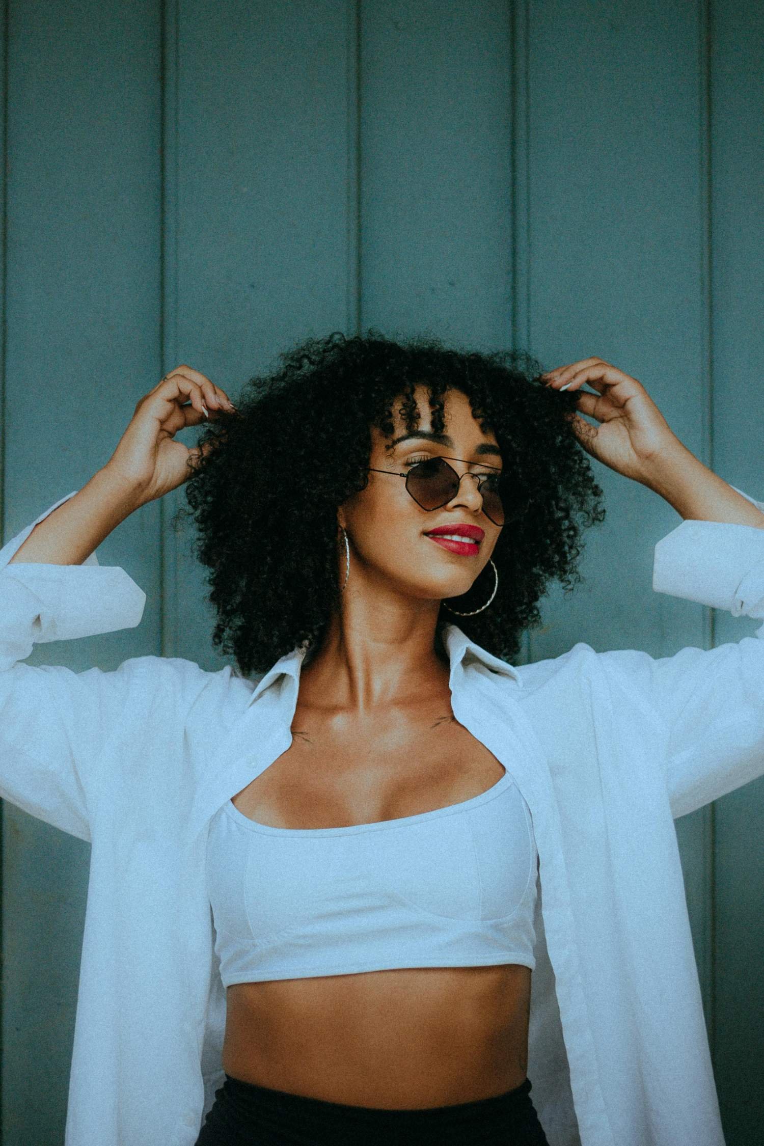 A woman with curly hair wearing sunglasses, white shirt, and earrings, posing with her hands on her head against a blue wall.