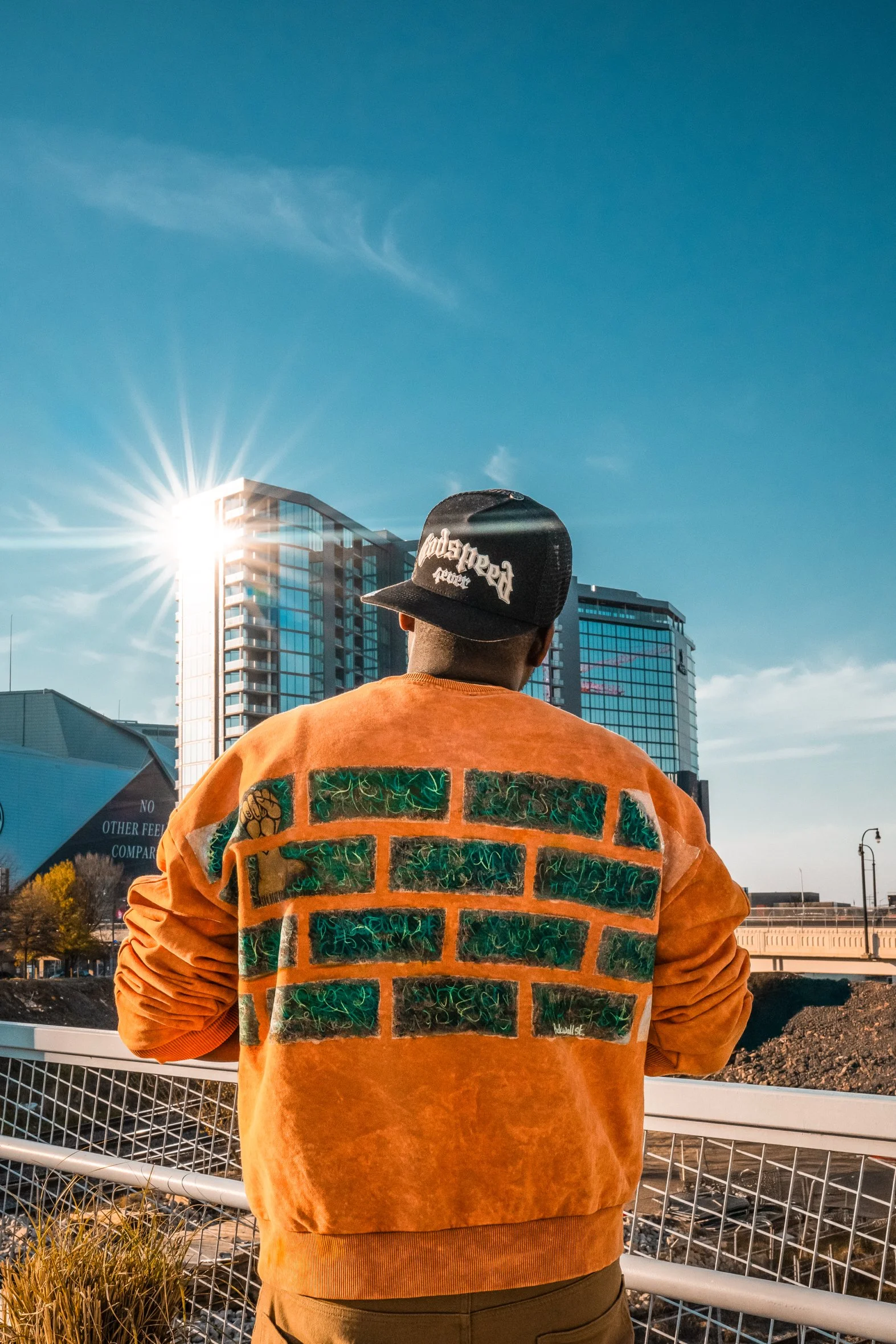 A person wearing an orange jacket with green and black graphic design on the back, a black cap with white embroidered text, facing away in an urban setting with tall buildings and a bright sun in the sky.