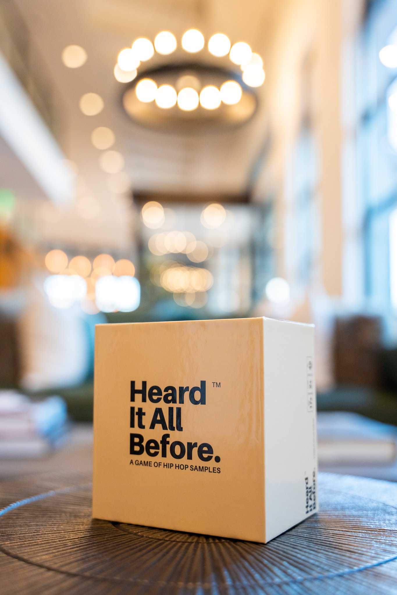 A cardboard box with the text "Heard It All Before" placed on a wooden table in a well-lit indoor space with blurred hanging lights and windows in the background.