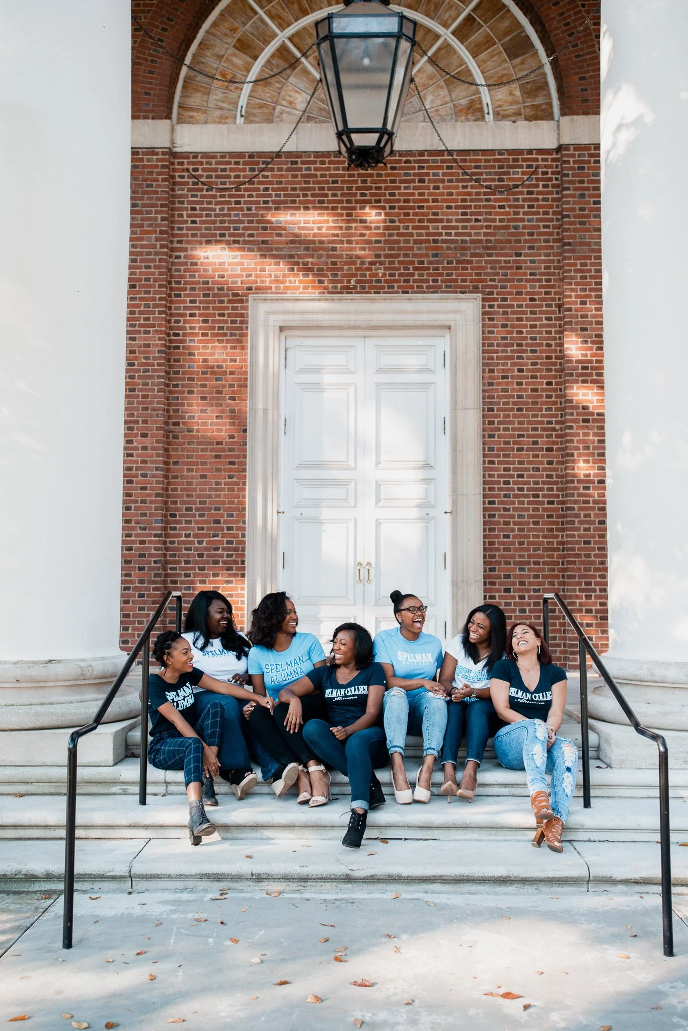 A group of young women sitting on steps in front of a large brick building with white double doors, laughing and talking together, wearing matching blue and black t-shirts that say 'SPELMAN ALUMNA' and 'SPELMAN COLLEGE'.