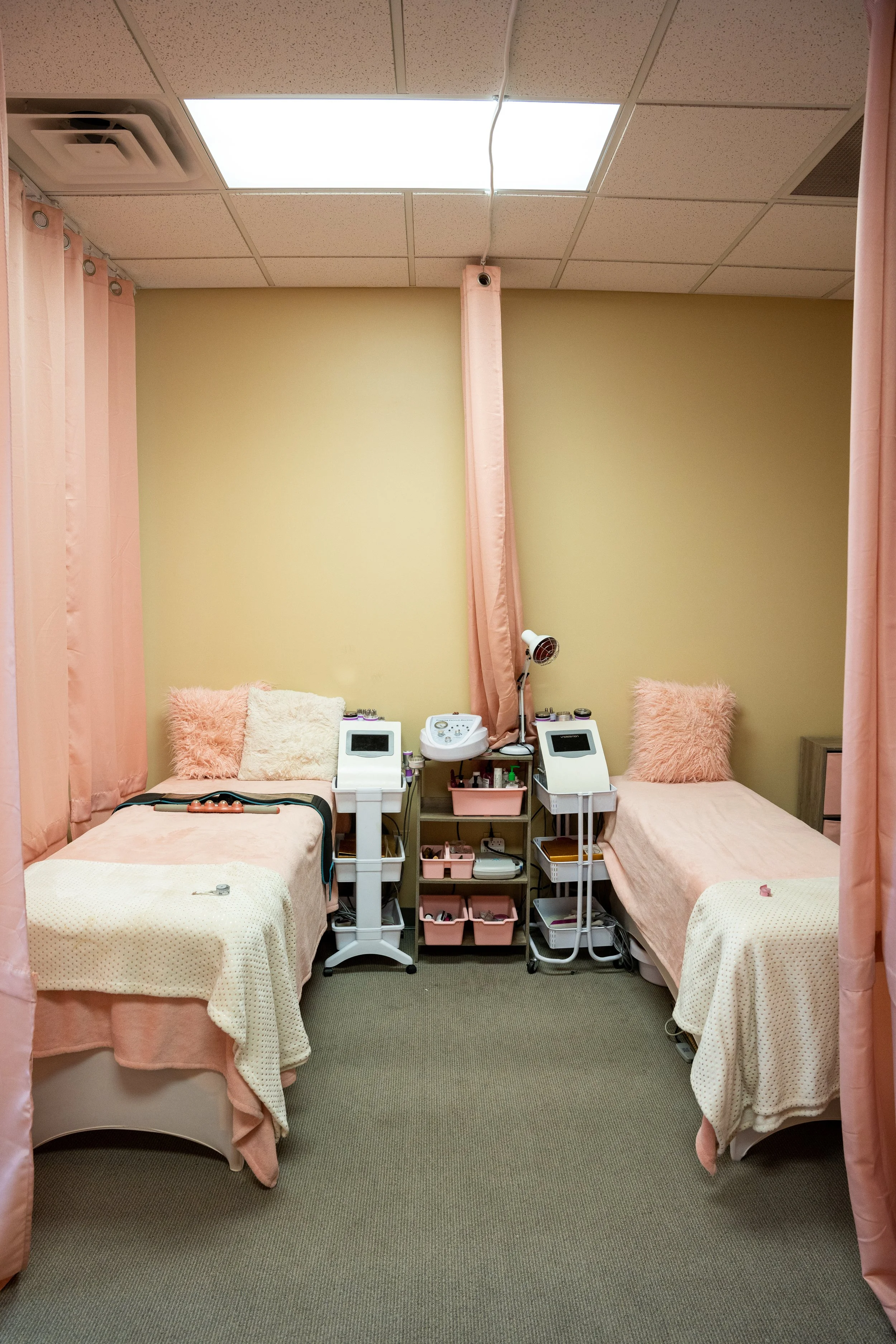Two medical treatment beds with pink pillows and beige blankets, separated by pink curtains. Medical equipment, including monitors and supplies, are positioned between the beds in a clinical room with beige walls and a ceiling with fluorescent lights.