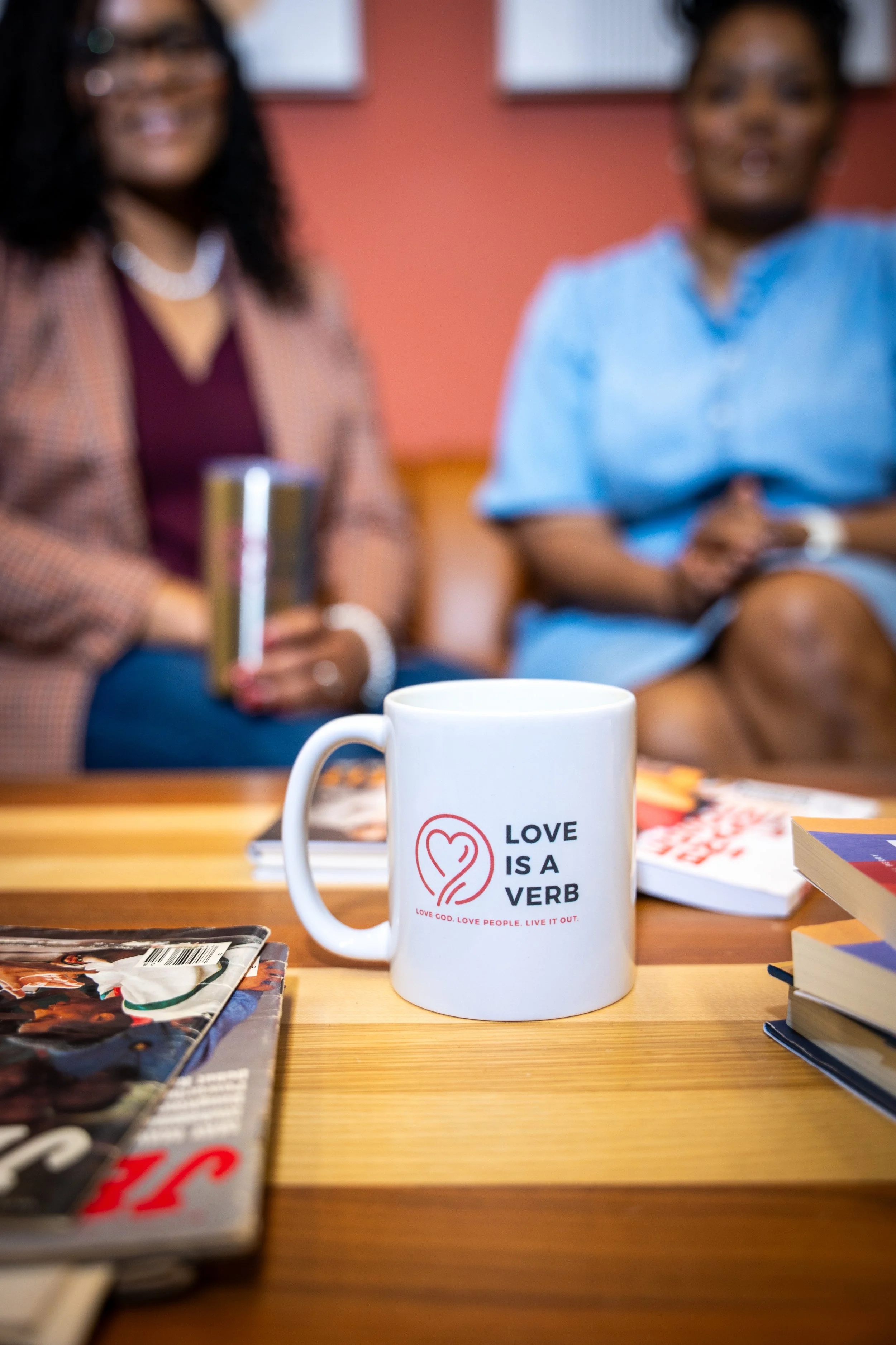 A white coffee mug with the text "Love is a Verb" and a heart-shaped logo sits on a wooden table, with magazines and books nearby. Two women are blurred in the background, sitting in a room with red walls and framed pictures.