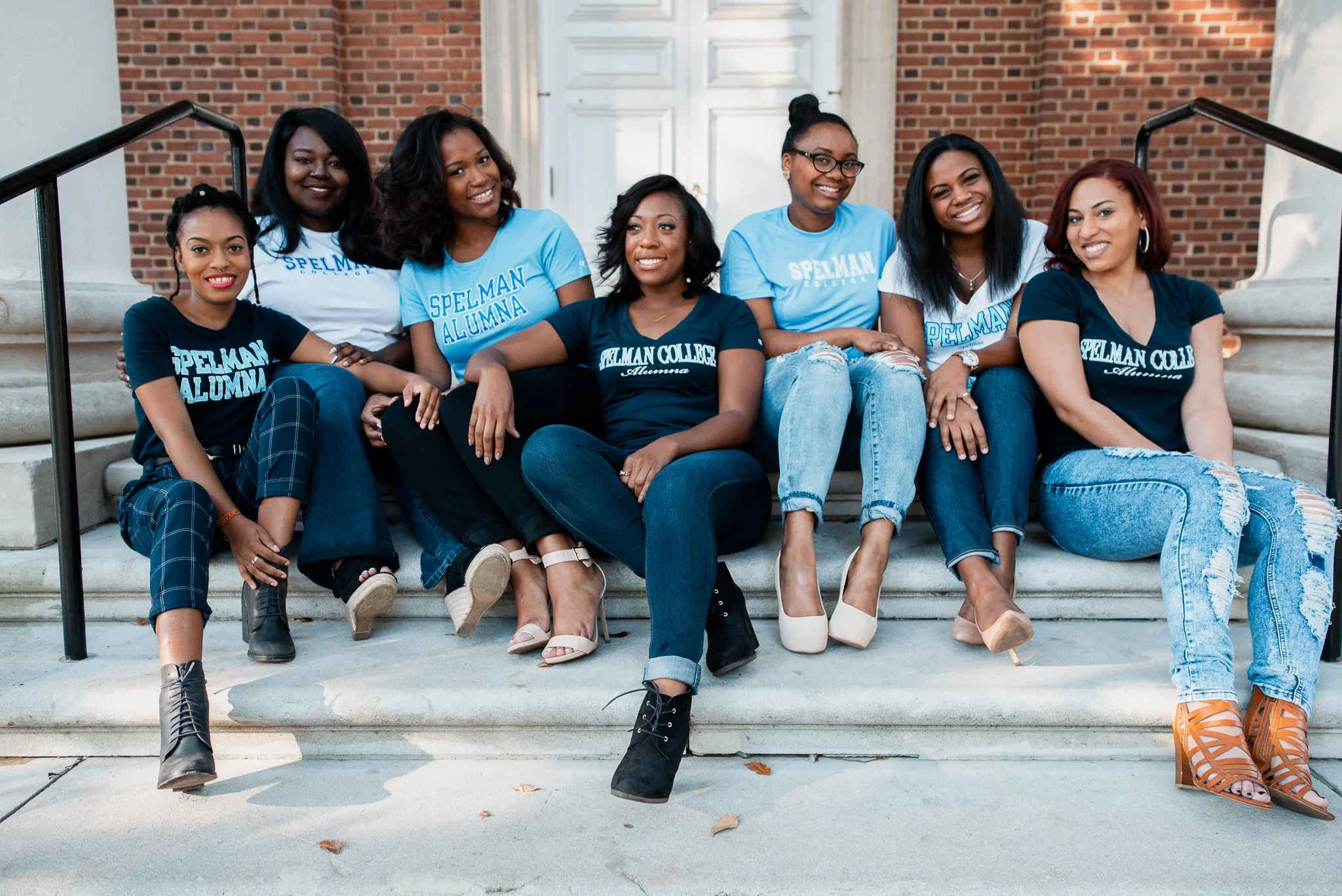 Group of seven women sitting on steps in front of a brick building, smiling at the camera. They are wearing shirts with 'Spelman College Alumna' written on them.