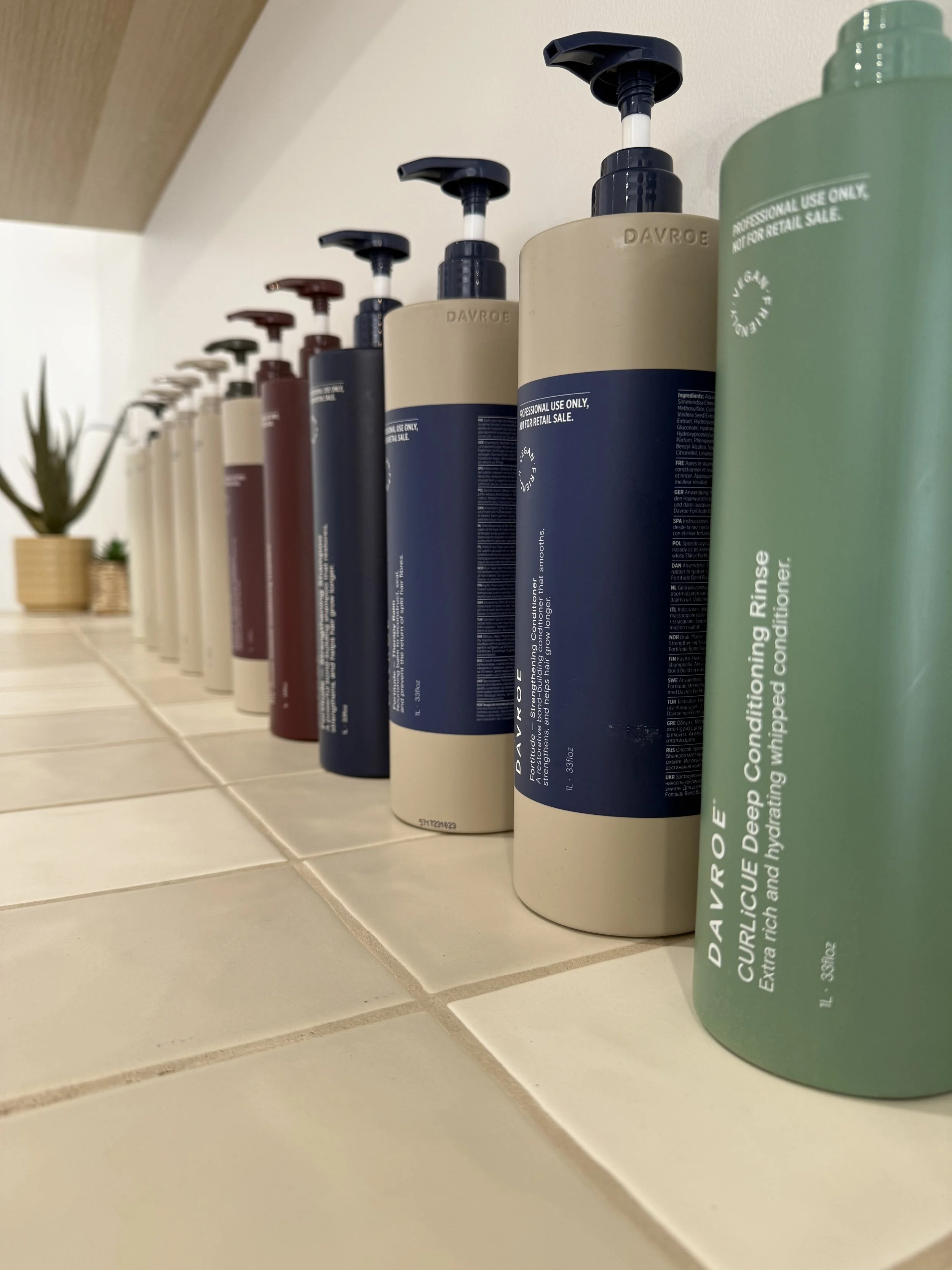 A row of shampoo and conditioner bottles on a bathroom tile shelf. The bottles are of different colors and sizes, with some having pump dispensers. In the background, there is a small potted plant.
