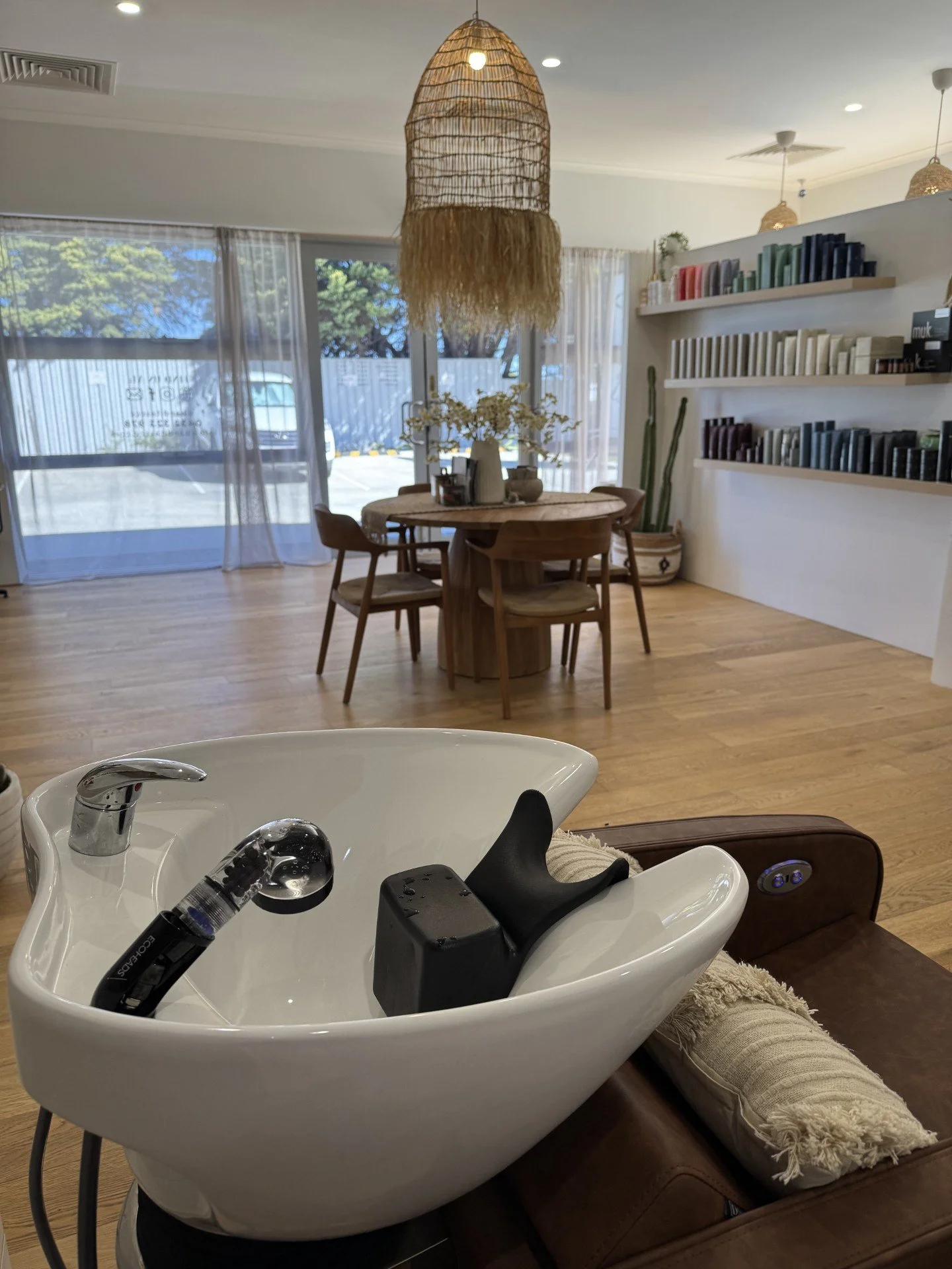 Indoor scene at a salon with a white shampoo bowl, a black shampoo bottle, and a hose for washing hair in the foreground. In the background, there is a round wooden table with chairs, a large window with curtains, and wall shelves with books and decorative items.