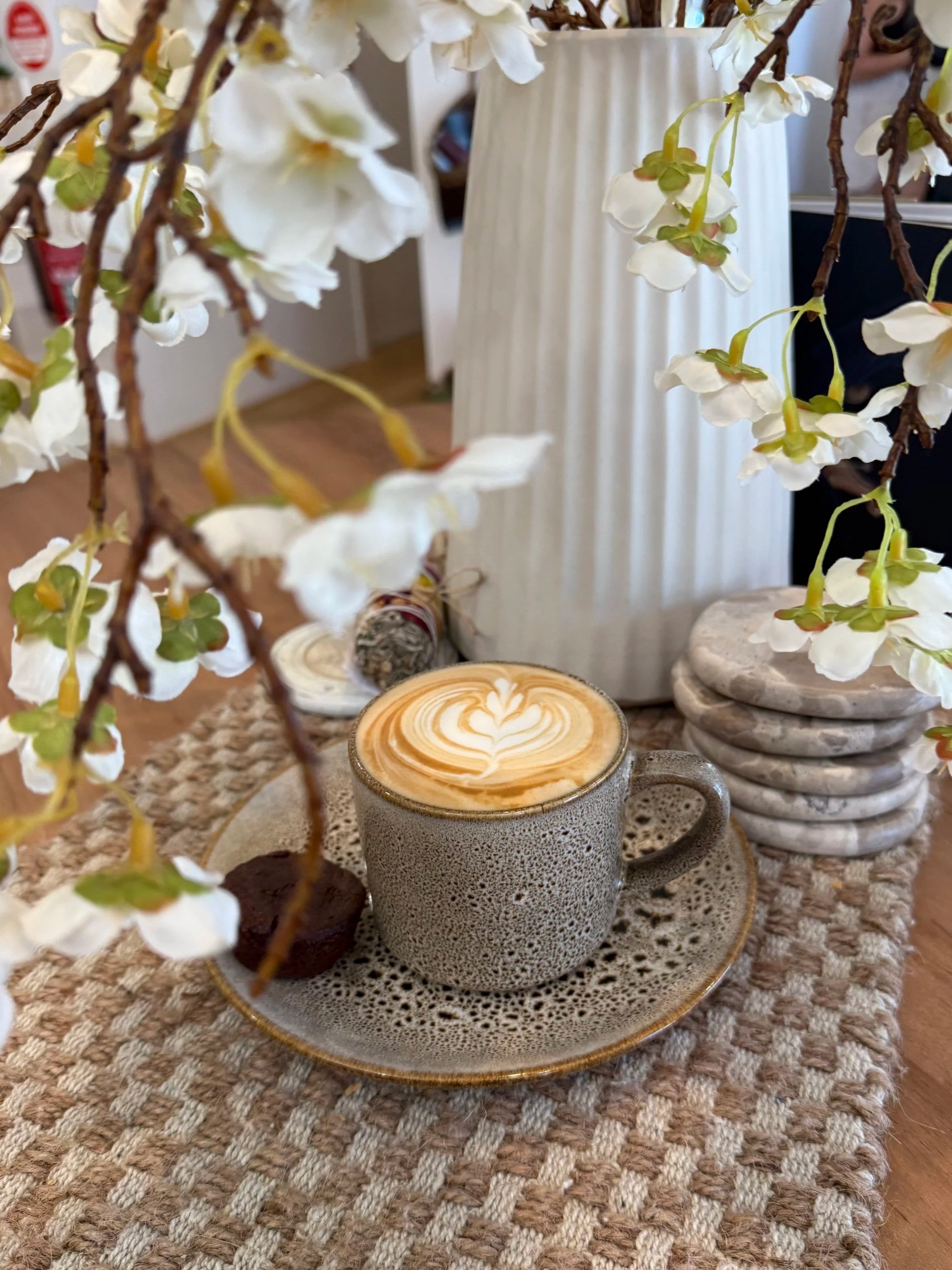 A ceramic cup of coffee with foam art on a woven tablecloth, surrounded by white flowers in a vase and decorative stones.