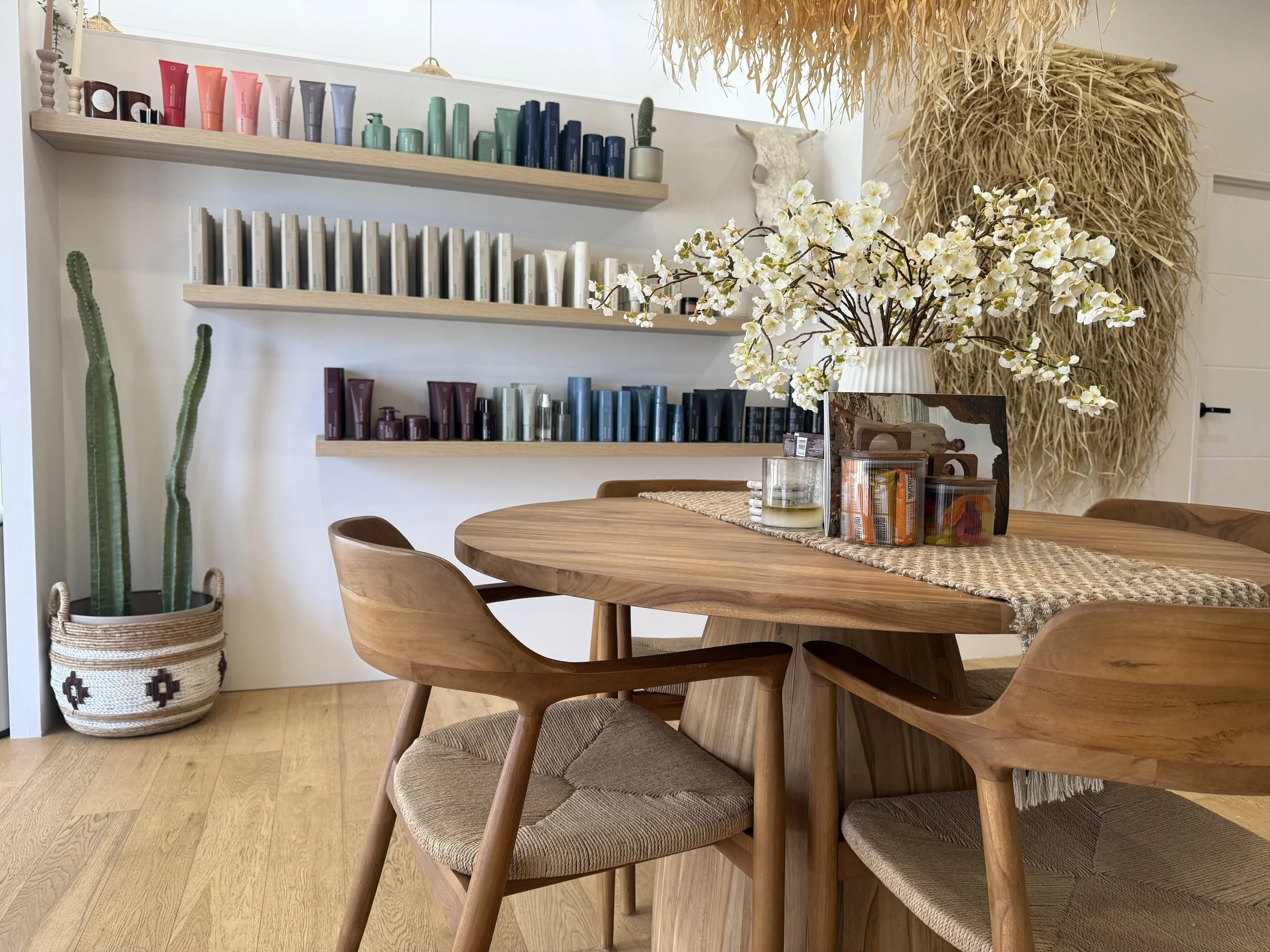 A wooden dining table with a woven table runner, a white flower arrangement, and a clear glass of water with a straw in a modern dining room. Behind the table, there are wooden chairs with woven seats, a set of three wooden shelves with skincare products, and green cacti in a basket. Decor includes a skull wall-mounted and a large dried plant or grass arrangement on the wall.