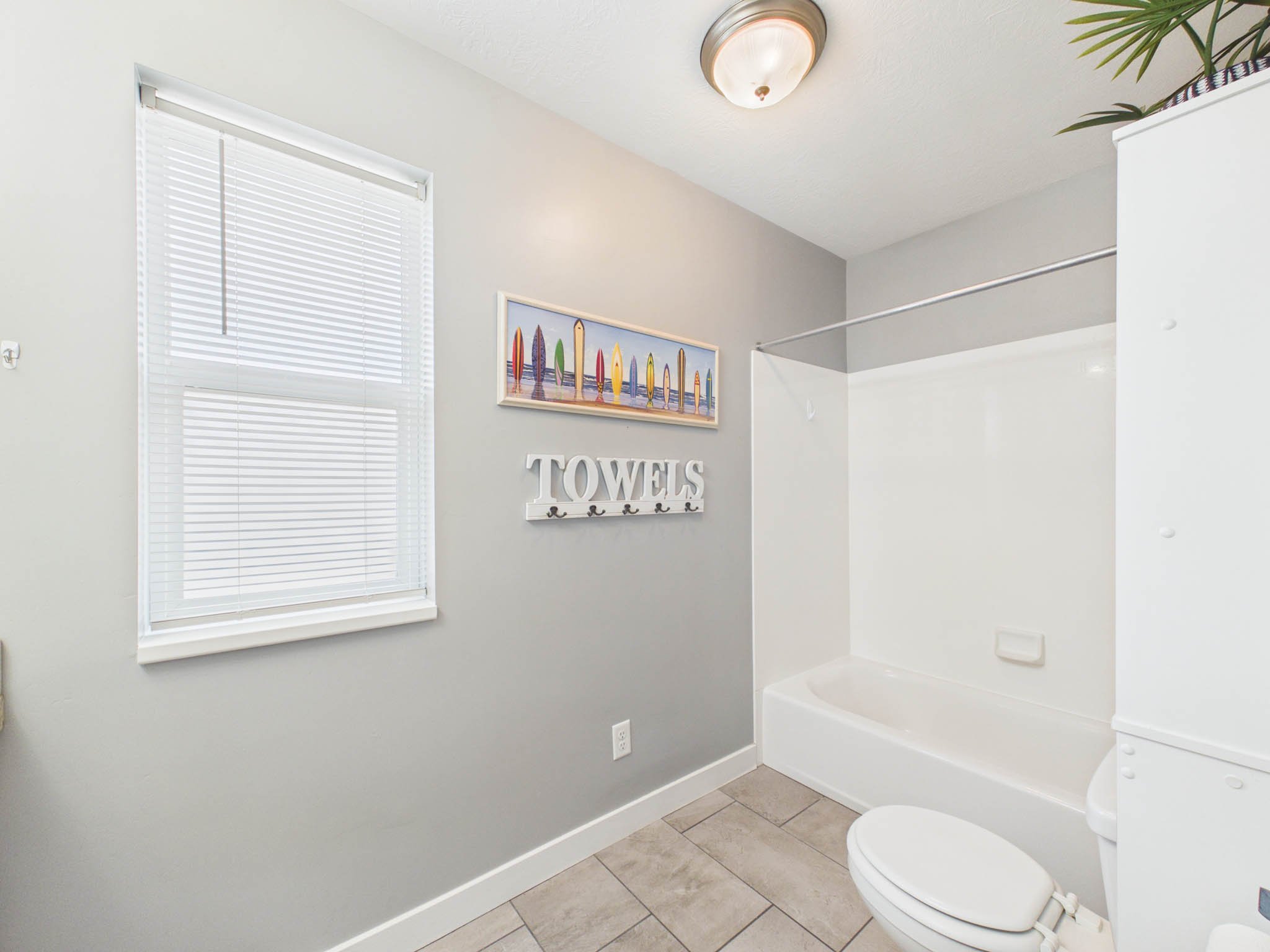 A bathroom with a window with white blinds, gray walls, a framed picture of colorful surfboards, a white towel rack sign, a toilet, and a bathtub.
