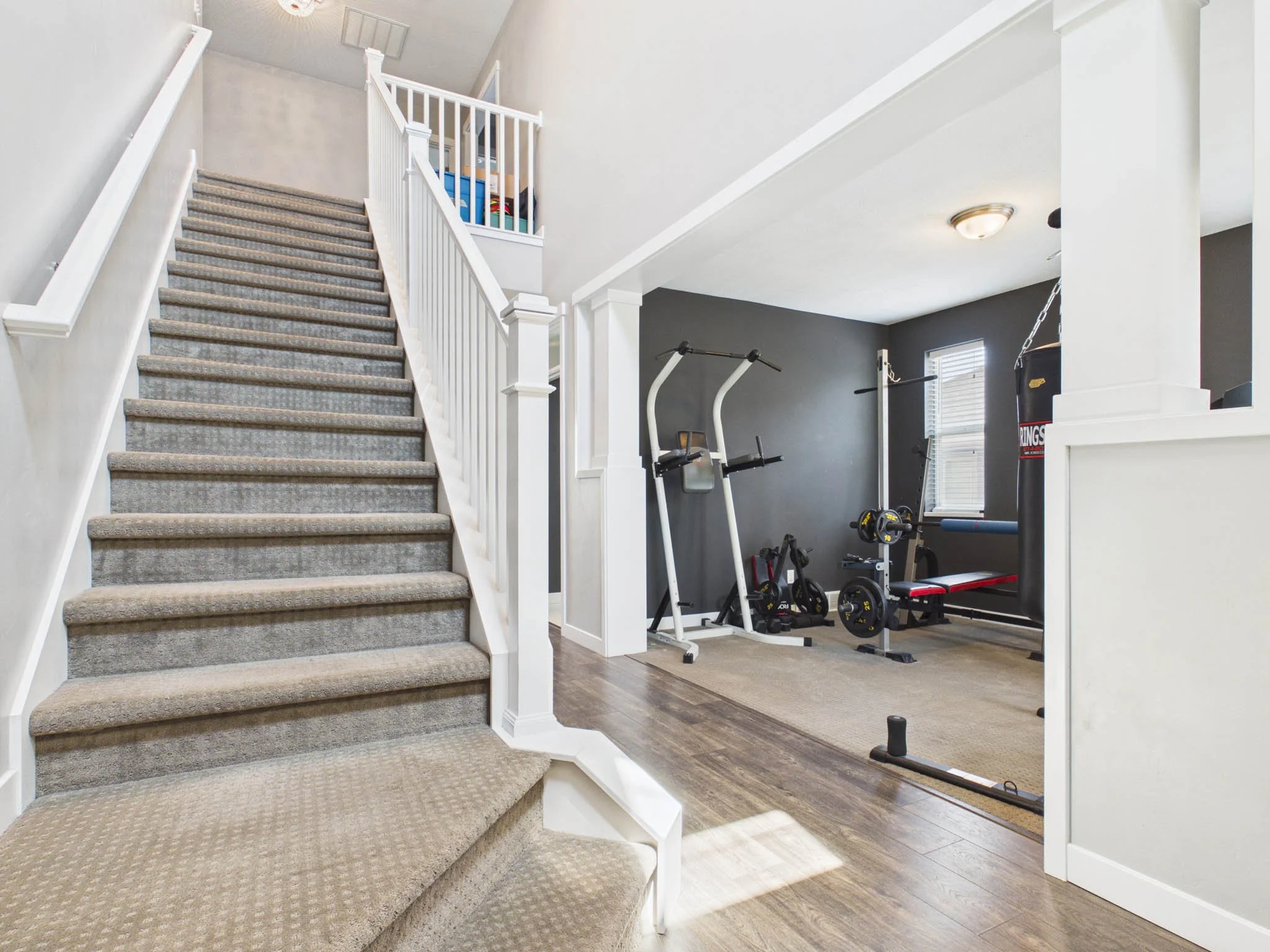 Interior view of a home gym area with carpeted stairs leading upstairs, workout equipment including weights and a pull-up bar in a room with dark gray walls, a window, and a light fixture on the ceiling.