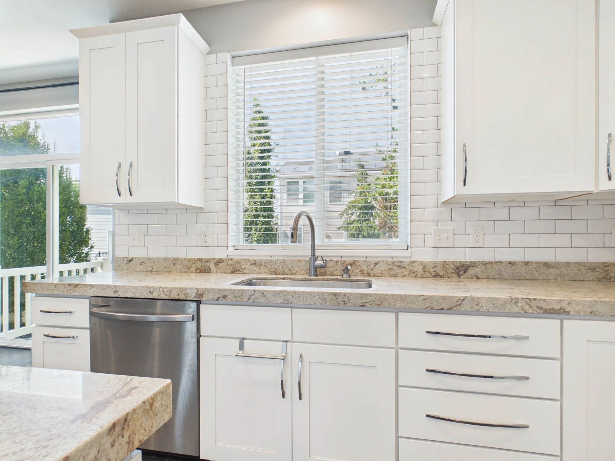 White kitchen cabinets with silver handles, a granite countertop, and a large window with white blinds showing trees outside.