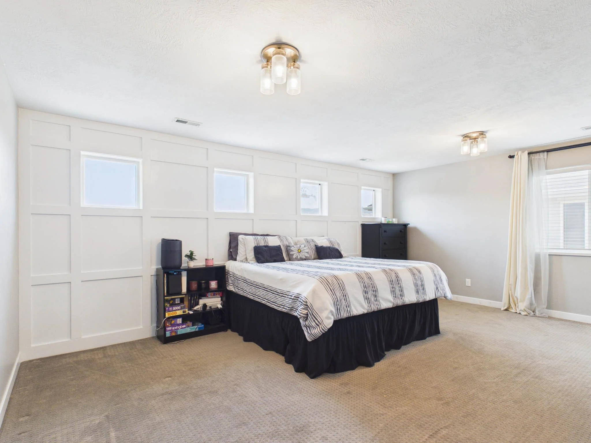 A bedroom with a bed covered in striped bedding, a small black nightstand with books and decor, white paneled walls, multiple windows, cream-colored carpet, and ceiling lights.