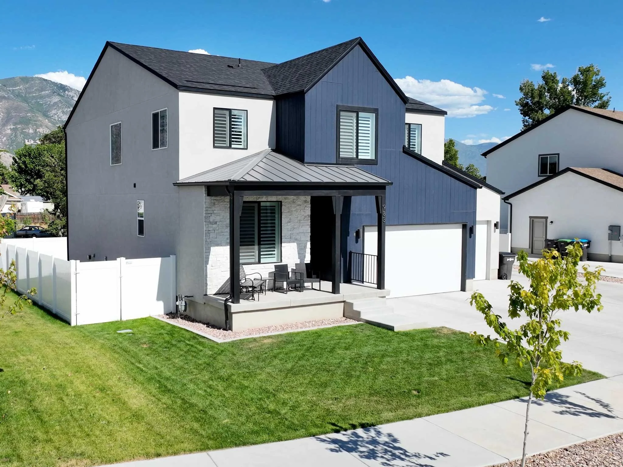 Modern two-story house with white and dark blue exterior, front porch with chairs, trimmed lawn, and neighboring houses, mountain in background under blue sky.
