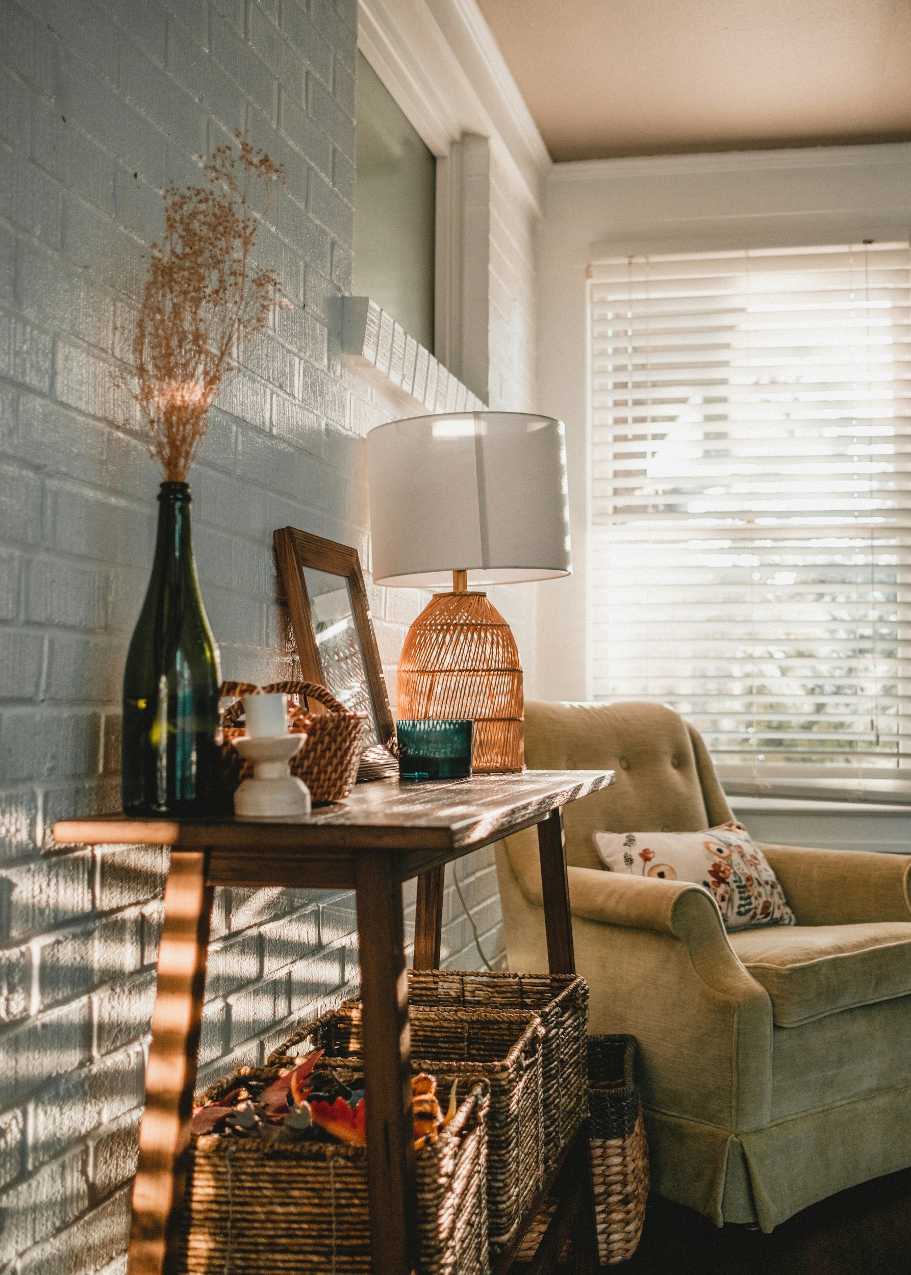 Interior of a cozy living room with a white brick wall, a wooden table with decorative items including a tall green vase with dried flowers, a candleholder, and a framed picture, a wicker table underneath, a beige sofa with a floral pillow, a white lamp with a woven base, and windows with horizontal blinds allowing sunlight to brighten the room.