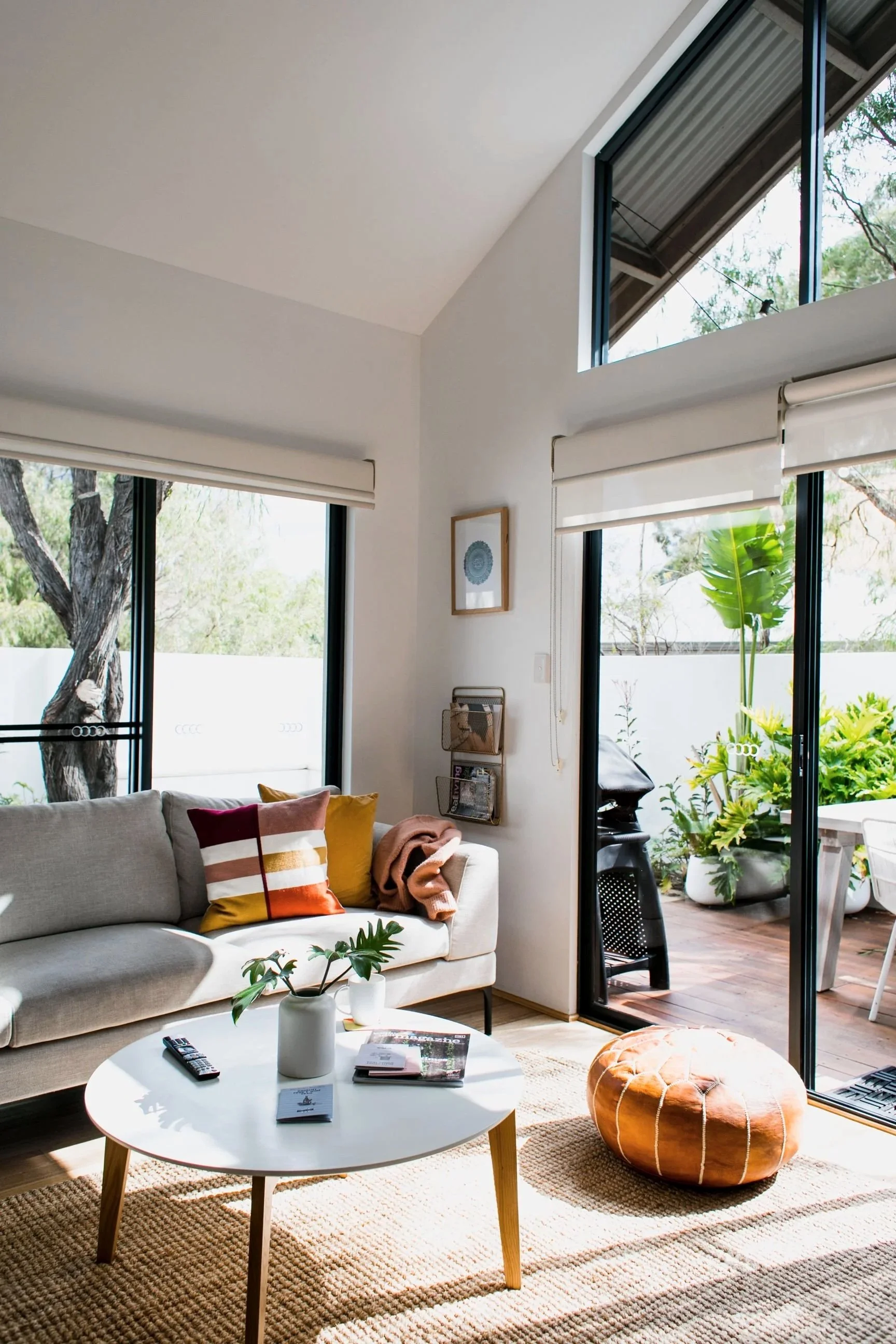 Living room with a beige sofa, pillow, coffee table with remote control and magazines, large windows with blinds, and sliding glass door leading to a patio with plants and outdoor furniture.