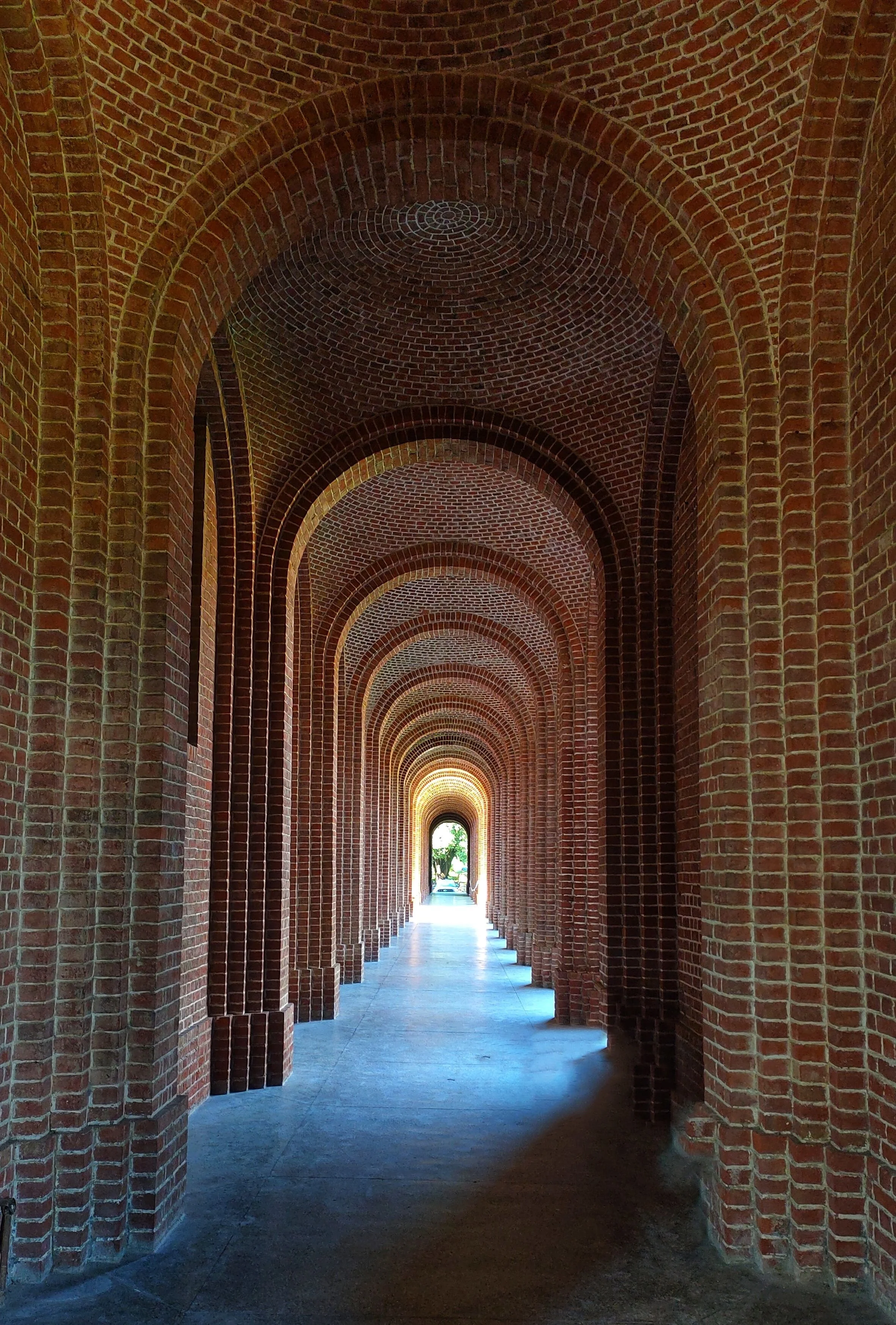 A long brick corridor with arched ceilings and doorways, leading to a bright light at the end.