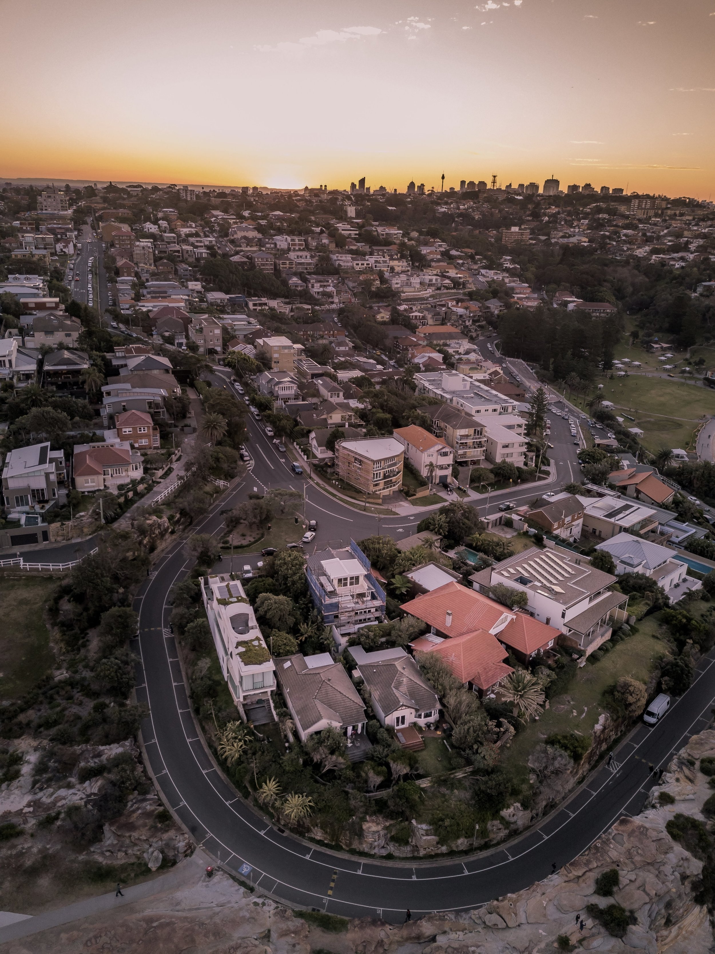 Aerial view of a residential neighborhood at sunset with houses, trees, and winding roads, overlooking a city skyline in the distance.