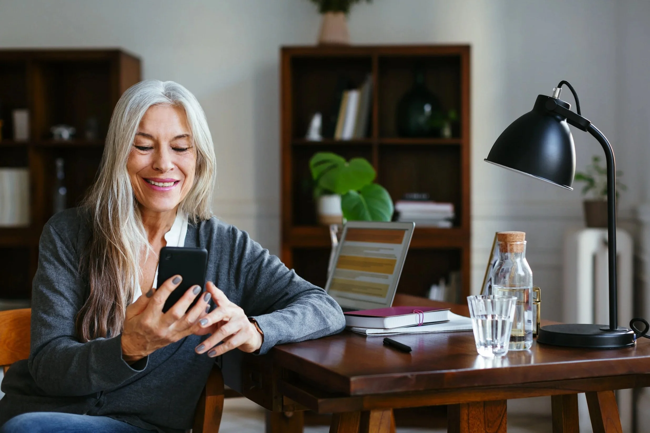 An elderly woman with long gray hair smiling while looking at her smartphone in a home office or living room. There is a black desk lamp, a glass of water, and some books and decorative items on the wooden table. A bookshelf with books and a plant is in the background.