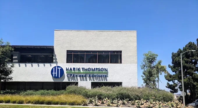The image shows a modern gray building with a sign that reads 'HARI K THOMPSON CPAs and Advisors,' featuring a blue circular logo with the initials 'HT'. The building is surrounded by greenery and a clear blue sky is in the background.